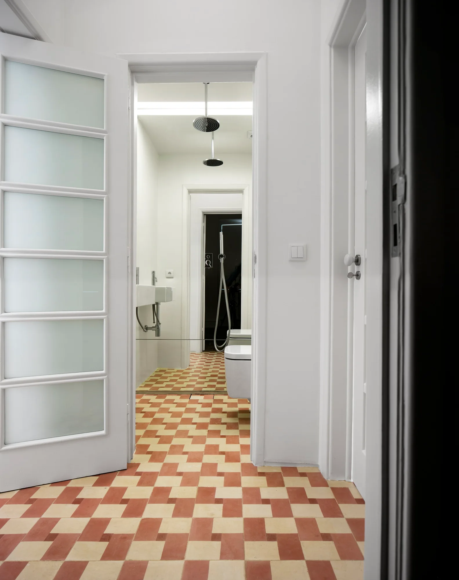 An open white door with frosted glass panels reveals a minimalist bathroom with terracotta and white checkered tile flooring.