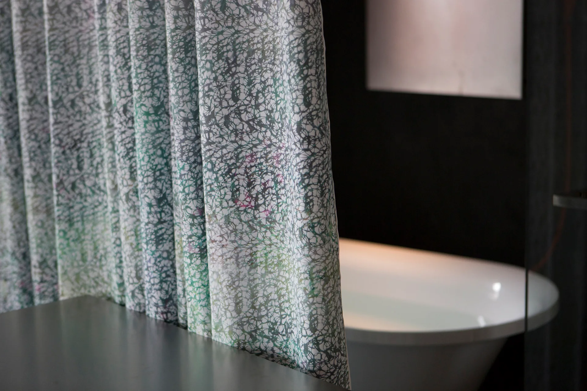 A green and white patterned fabric curtain partially blocks a white freestanding bathtub set against a dark matte wall in a bathroom.