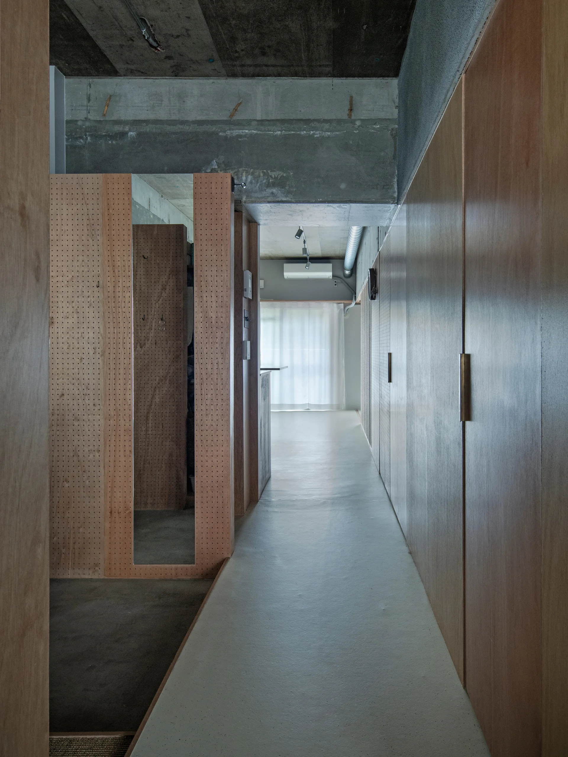A minimalist hallway featuring custom plywood built-in cabinets, a pegboard partition with a mirror, and an exposed concrete ceiling.