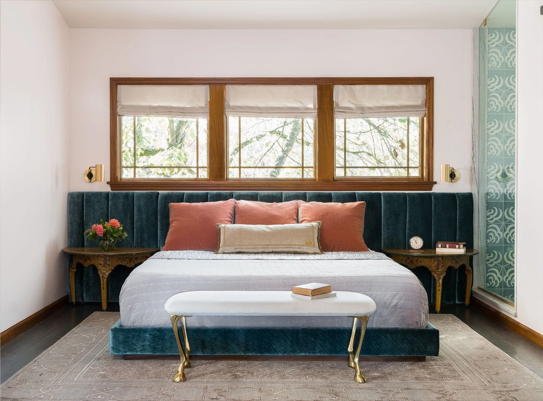 A bedroom features a teal velvet channel tufted headboard extending behind the bed and nightstands beneath a three panel wooden window.