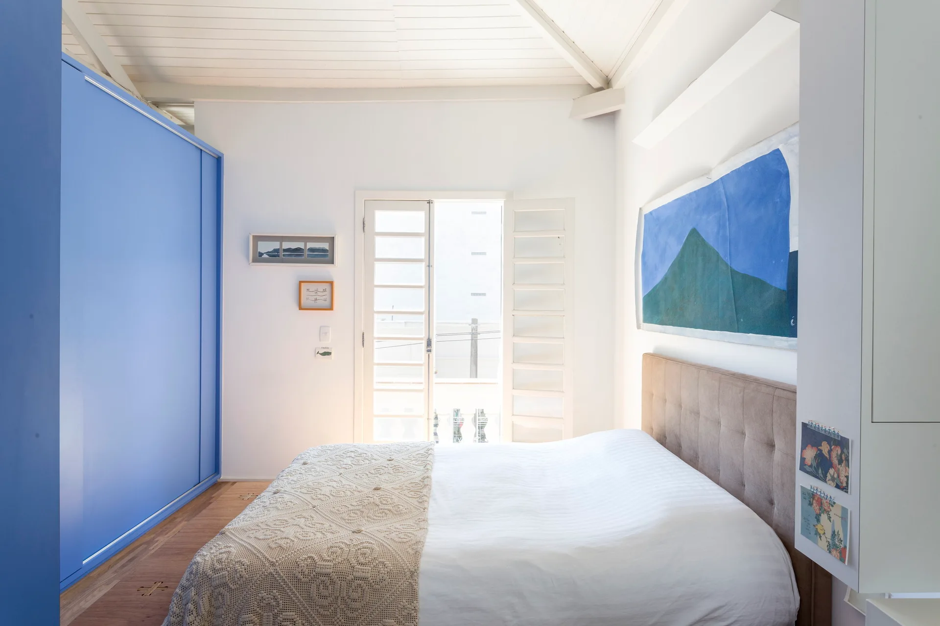 A bright bedroom features a bed with a tufted headboard, a large blue wardrobe, and a white planked ceiling with glass doors leading outside.