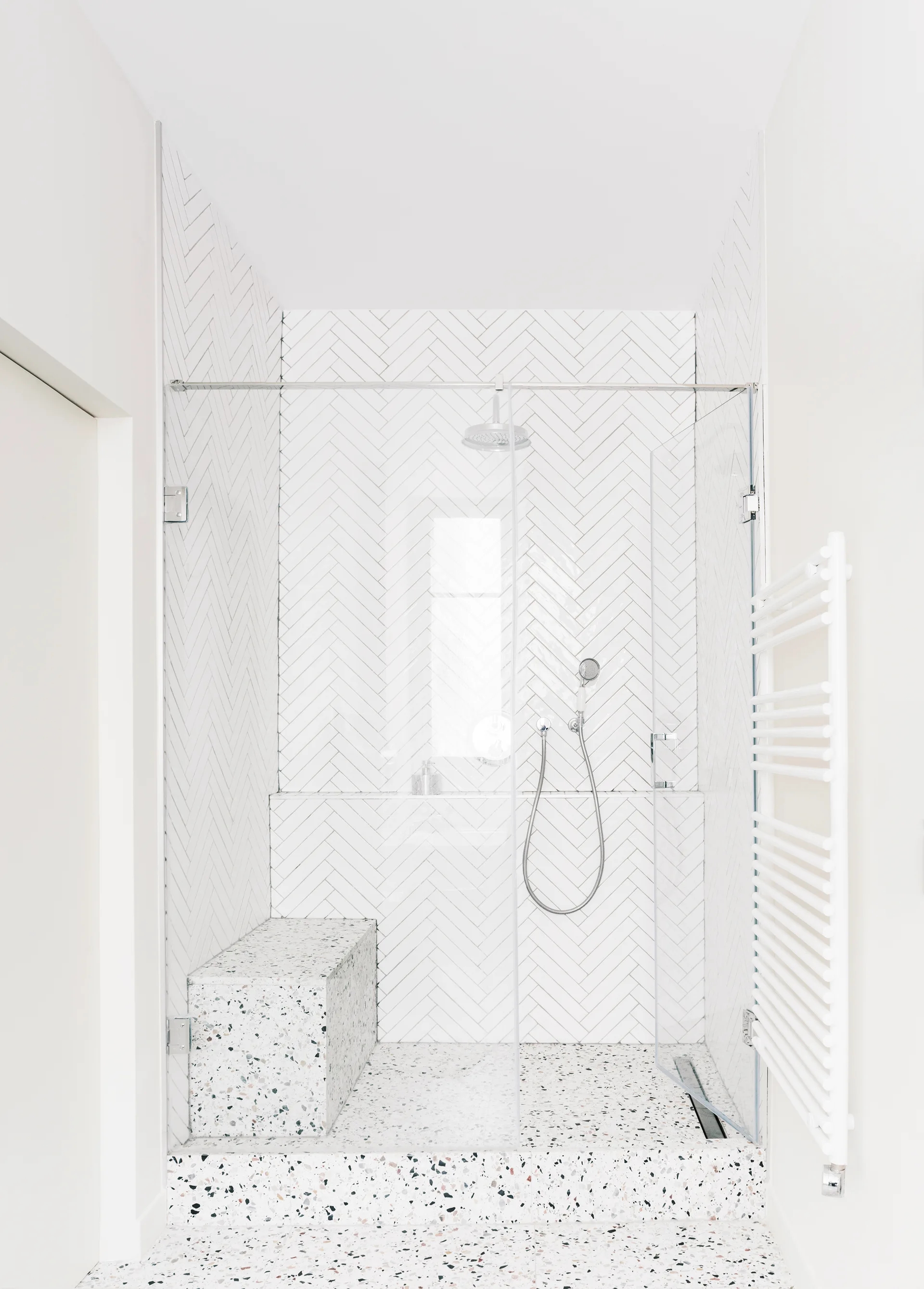 A modern walk-in shower features white herringbone pattern tiled walls, a glass enclosure, and a speckled terrazzo floor with a matching built-in bench.