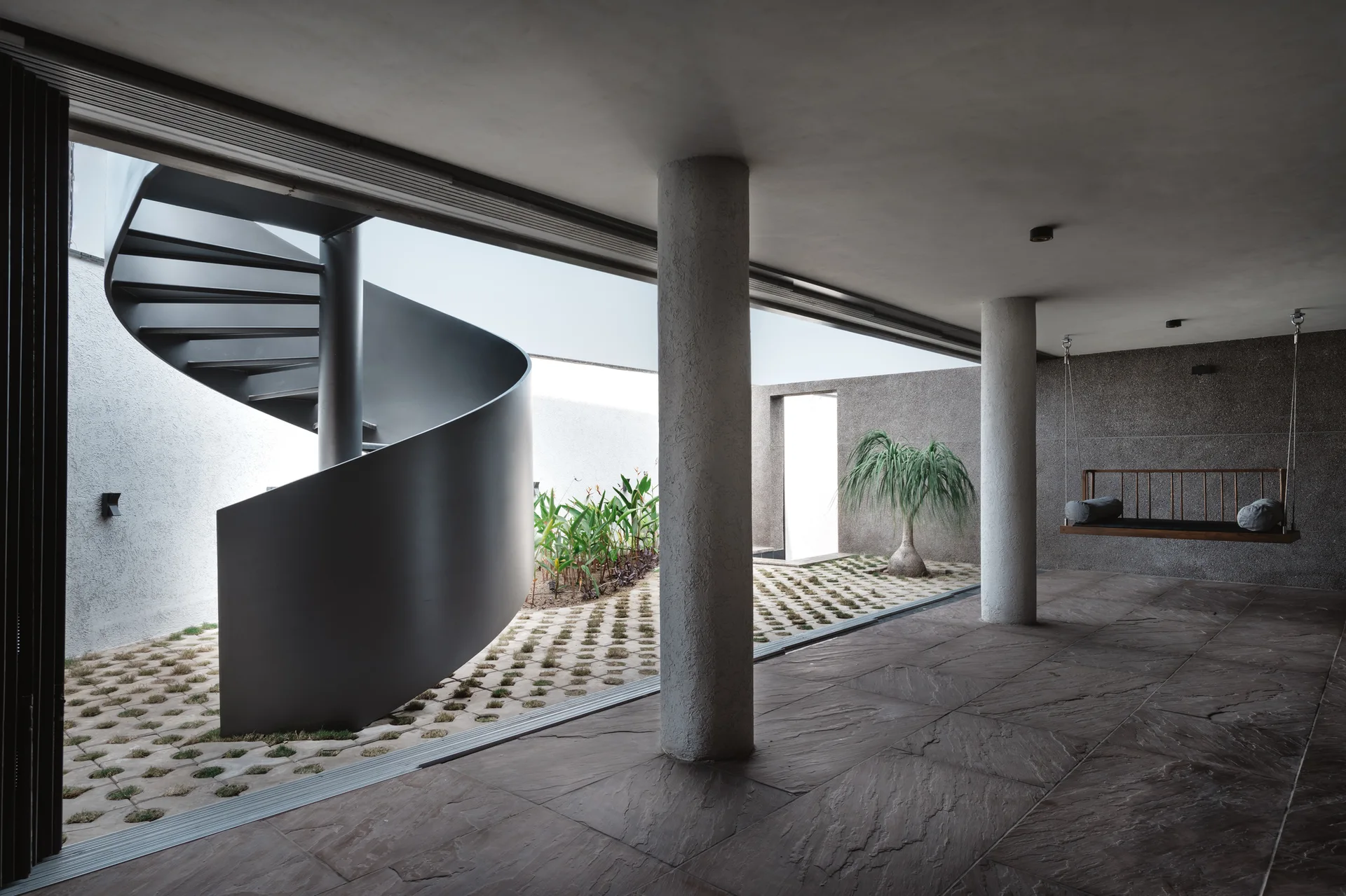 A covered concrete patio with stone floor tiles features a dark metal spiral staircase and a suspended wooden swing bench next to a paved courtyard.