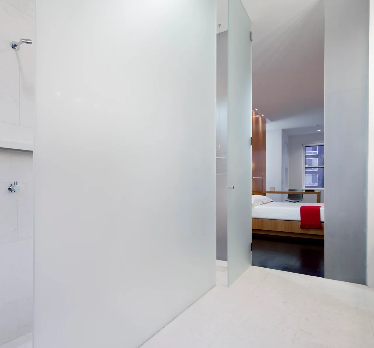 A view from a white-tiled shower area through a frosted glass door into a minimalist bedroom with a wooden bed frame and a dark floor.