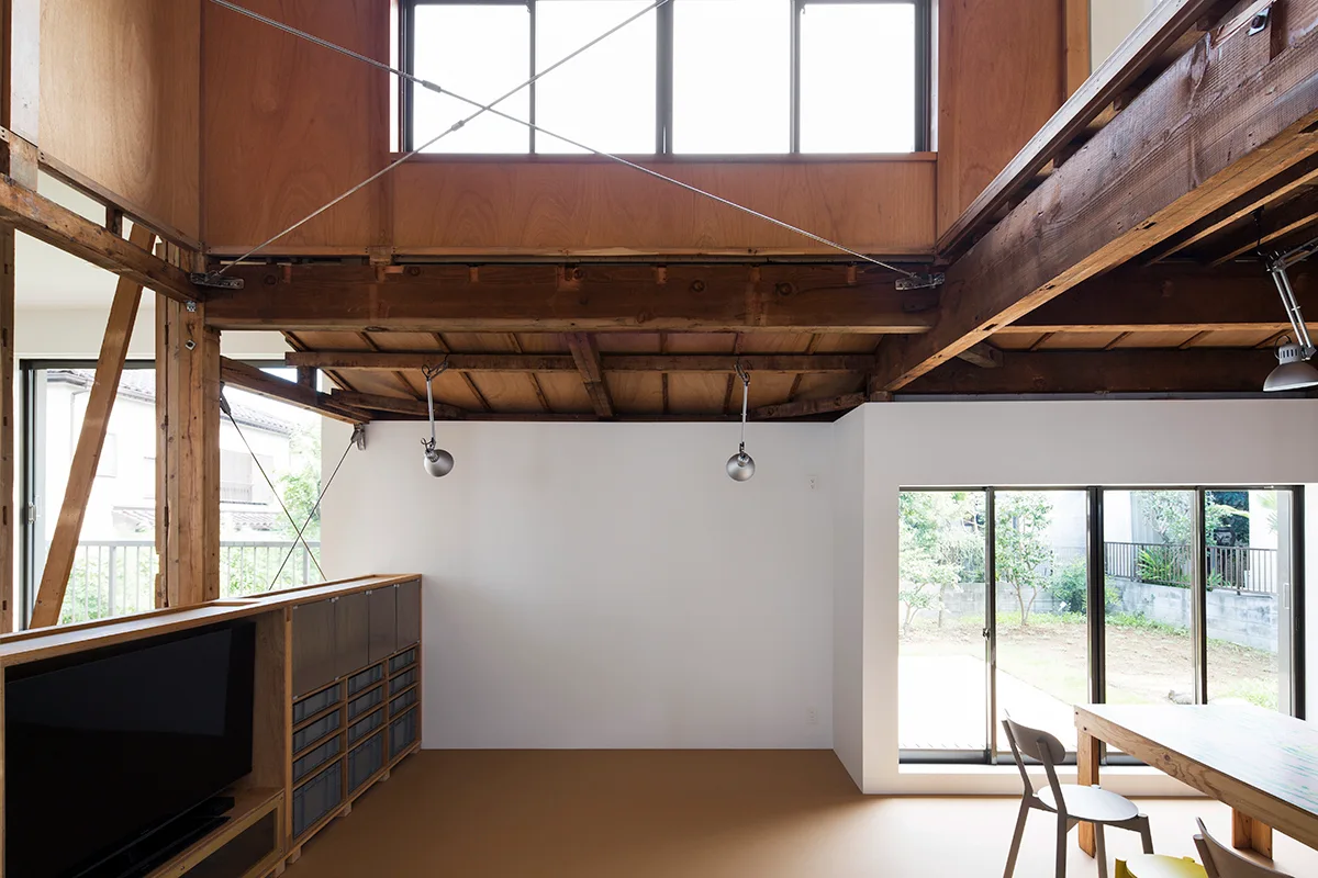 A double-height room featuring exposed dark timber beams, white walls, metal structural cables, and large glass doors looking onto a garden.