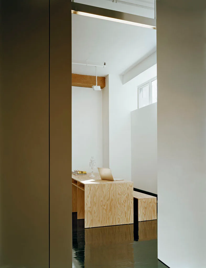 A minimalist home office featuring a light plywood desk and matching bench on a shiny black wood floor against white walls.