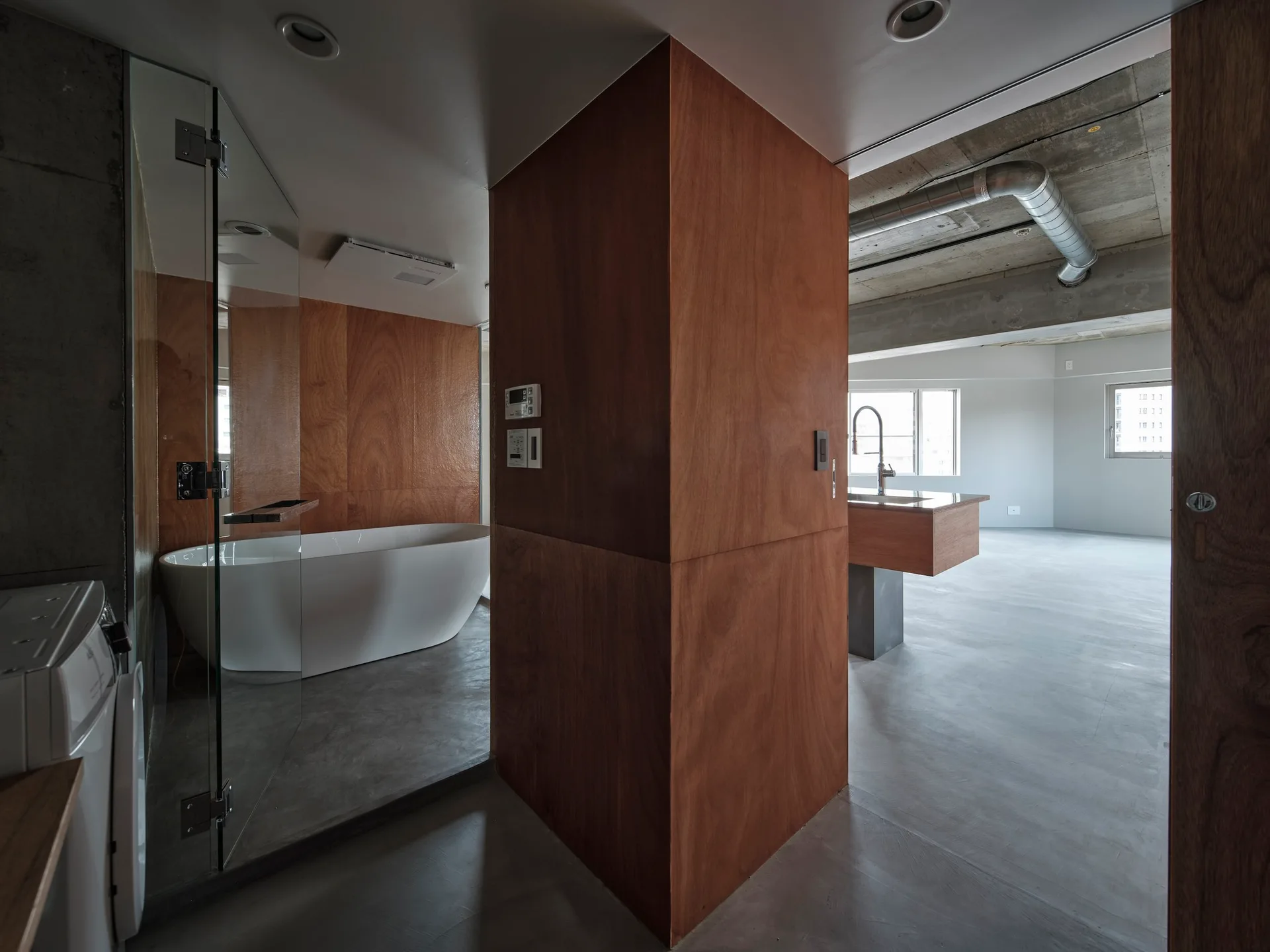 A modern open-concept loft features a glass-enclosed bathroom with a white freestanding tub next to a wood-clad pillar and concrete flooring.