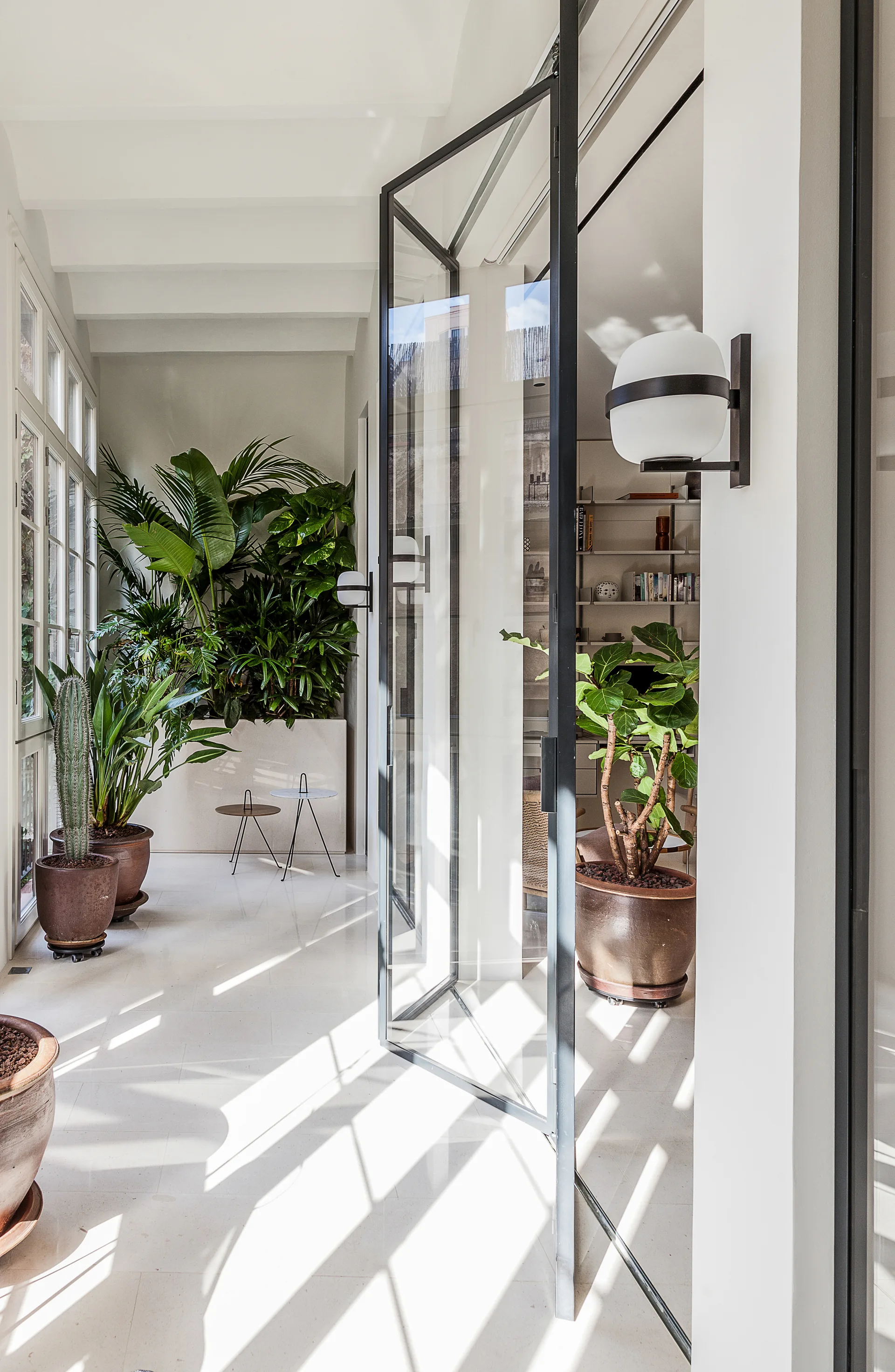 A sunlit white corridor features a large black-framed glass door, numerous potted tropical plants, and modern globe wall sconces.