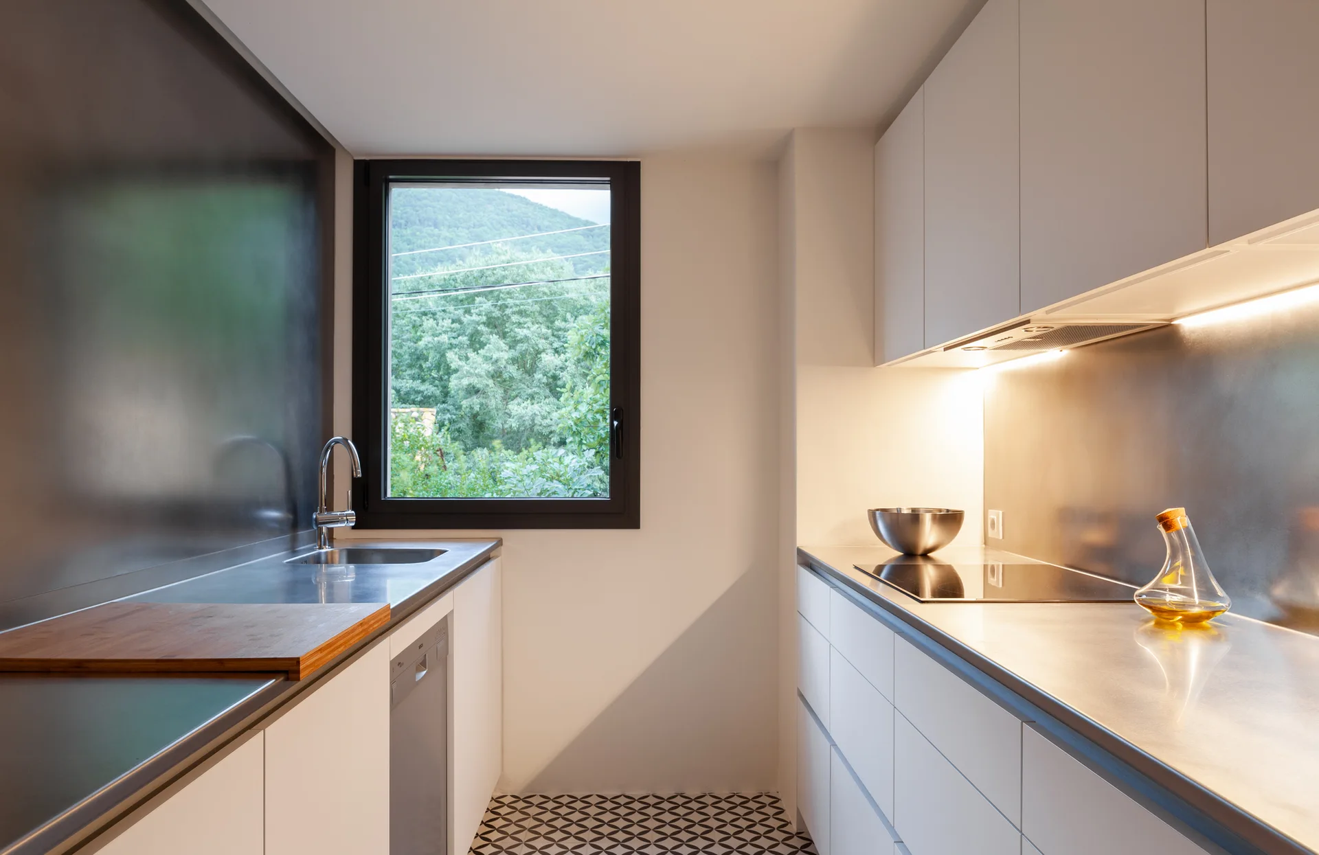 A galley kitchen featuring white handleless cabinets, stainless steel countertops, a black-framed window, and geometric black and white floor tiles.