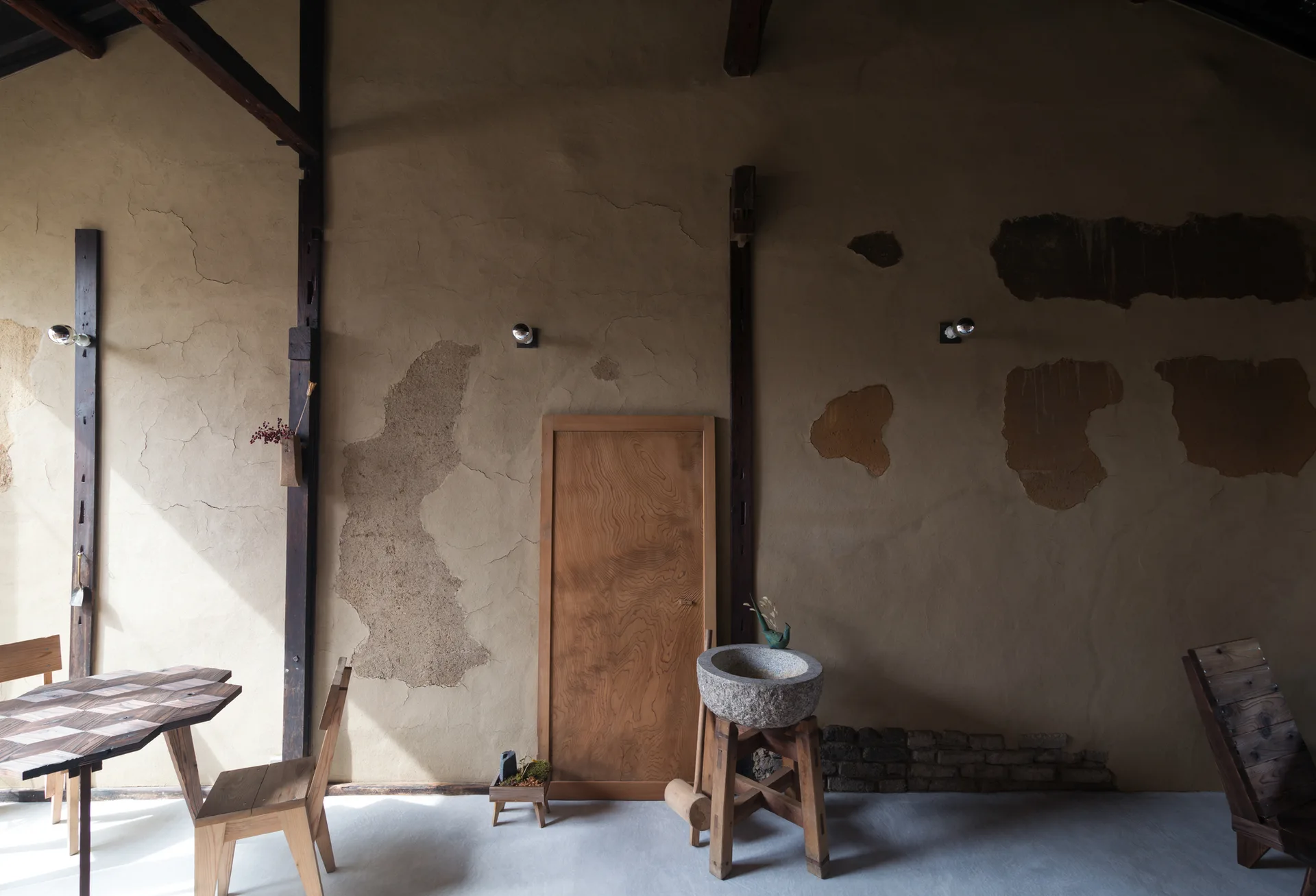 A rustic room features a textured beige plaster wall, a natural wood door, a stone basin on a stand, and a wooden table with chairs.