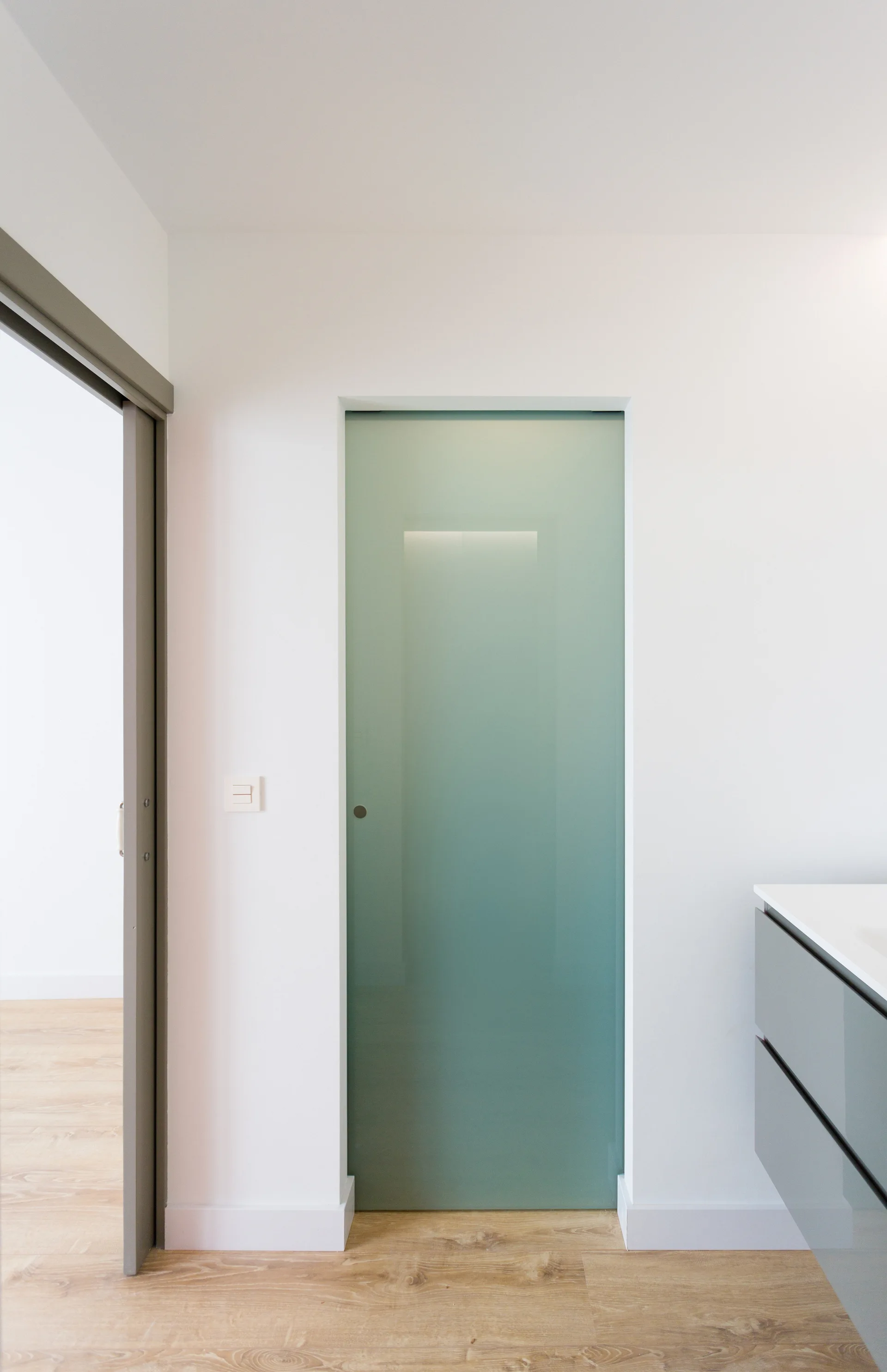 A frosted green glass sliding door is set into a plain white wall next to a glossy gray vanity cabinet in a minimalist room with wood flooring.