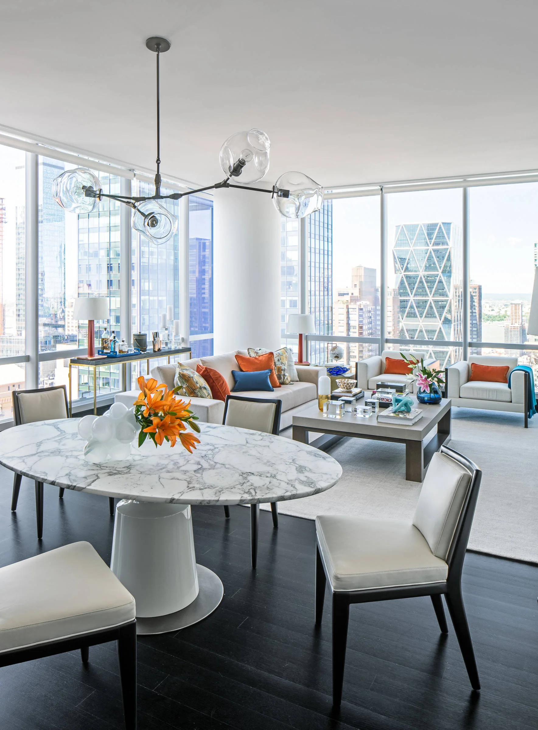 An open-concept living and dining area features an oval marble table, white chairs, a beige sofa, and floor-to-ceiling windows showing a city skyline.