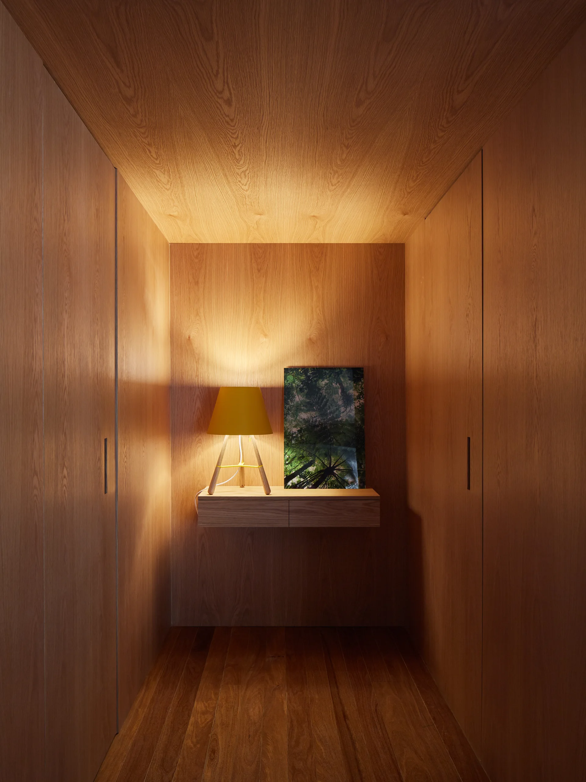A narrow walk-in closet fully lined in wood veneer with integrated wardrobes, a central floating shelf, a yellow lamp, and a framed print.