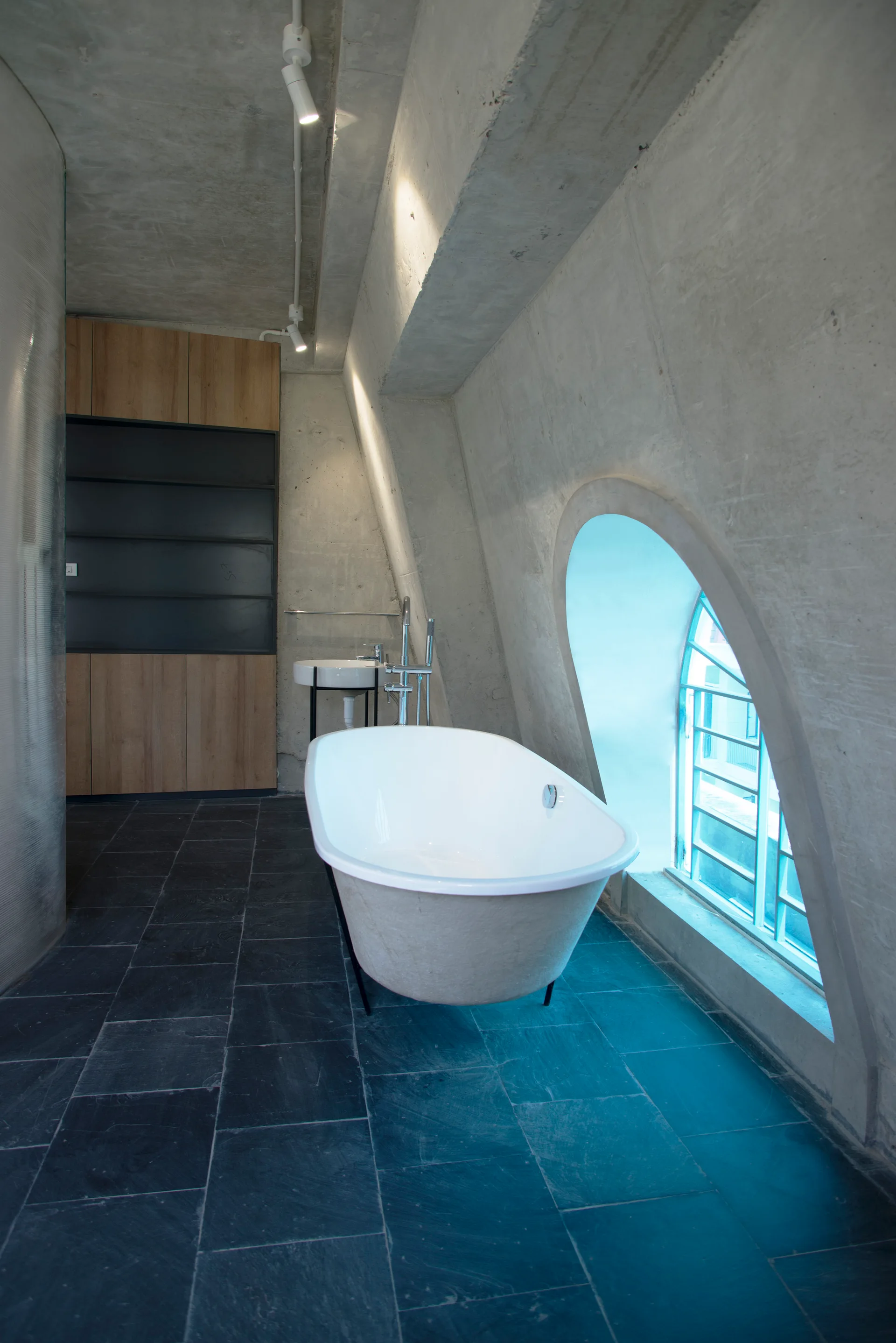 A modern industrial bathroom featuring a white freestanding bathtub on black tile flooring beside a sloped concrete wall with an arched window.