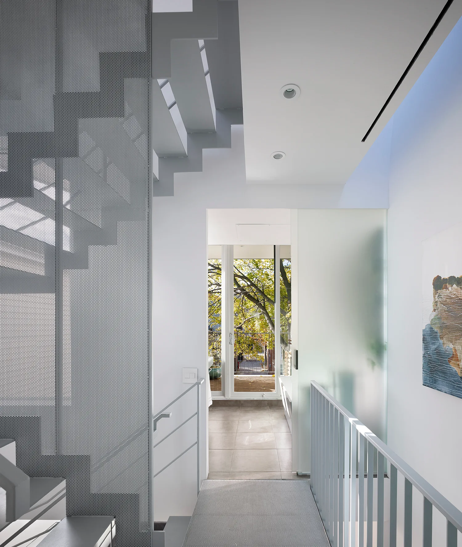 A modern hallway features a white staircase enclosed by a grey perforated metal screen, with a frosted glass sliding door leading to an outdoor view.