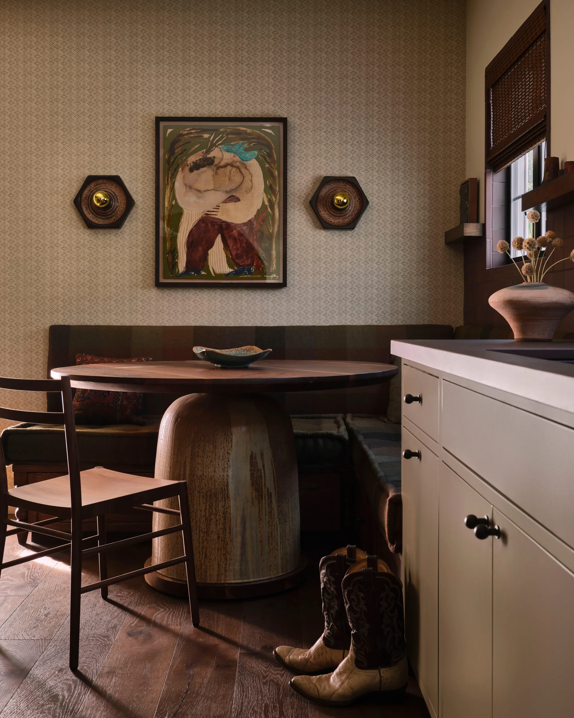 A cozy dining nook featuring a round wooden table, a corner built-in bench, patterned beige wallpaper, and a pair of cowboy boots on the wood floor.