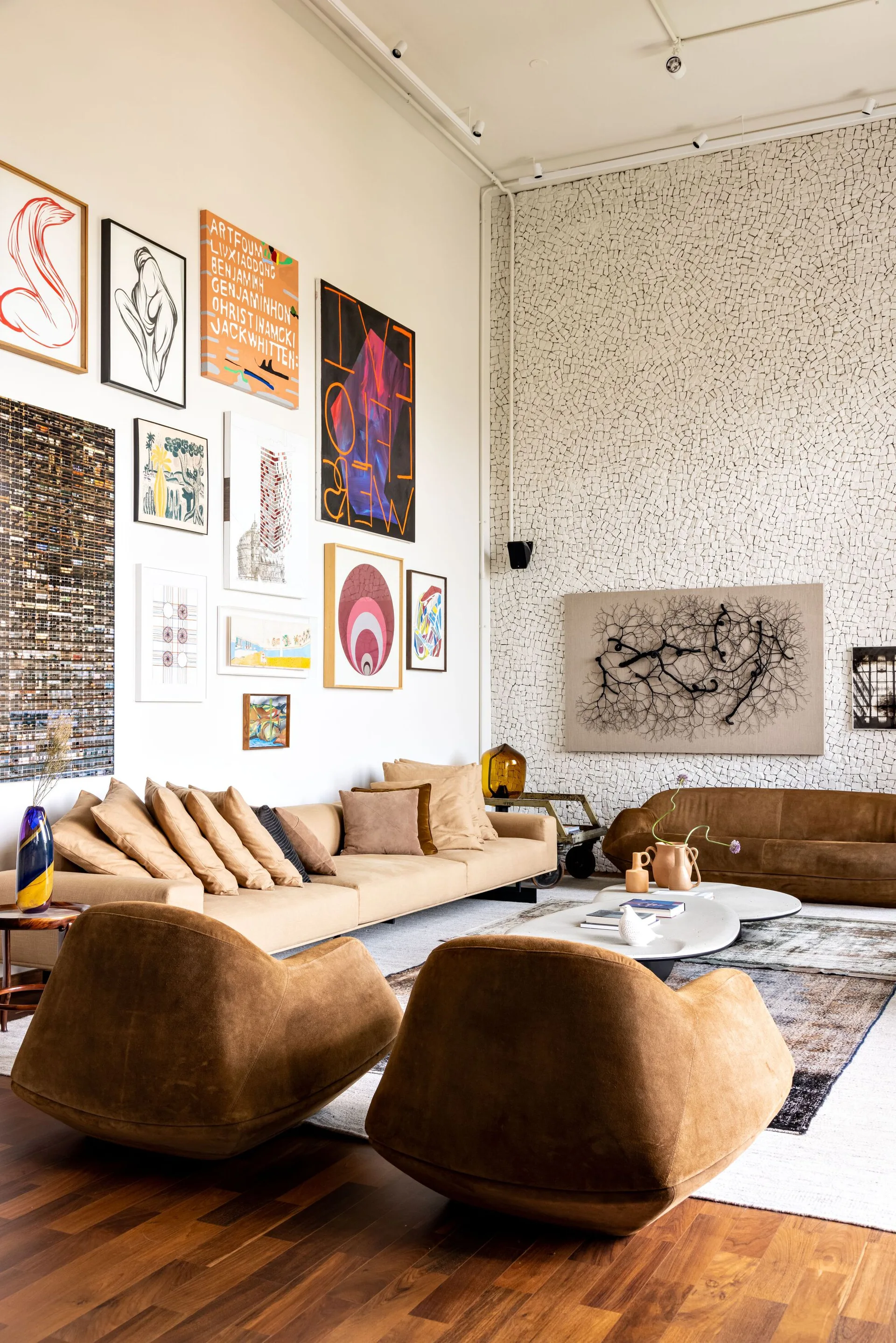 A spacious living room with low-profile tan armchairs, a beige sofa beneath a large gallery wall, and a textured white stone mosaic feature wall.