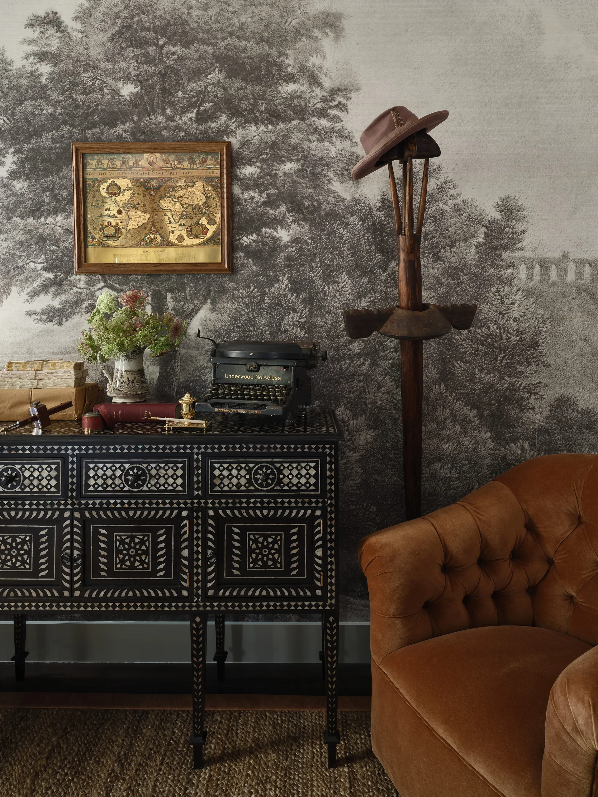 A black console table with intricate white geometric inlay is styled with vintage books, a typewriter, and an antique map against a forest mural wallpaper.