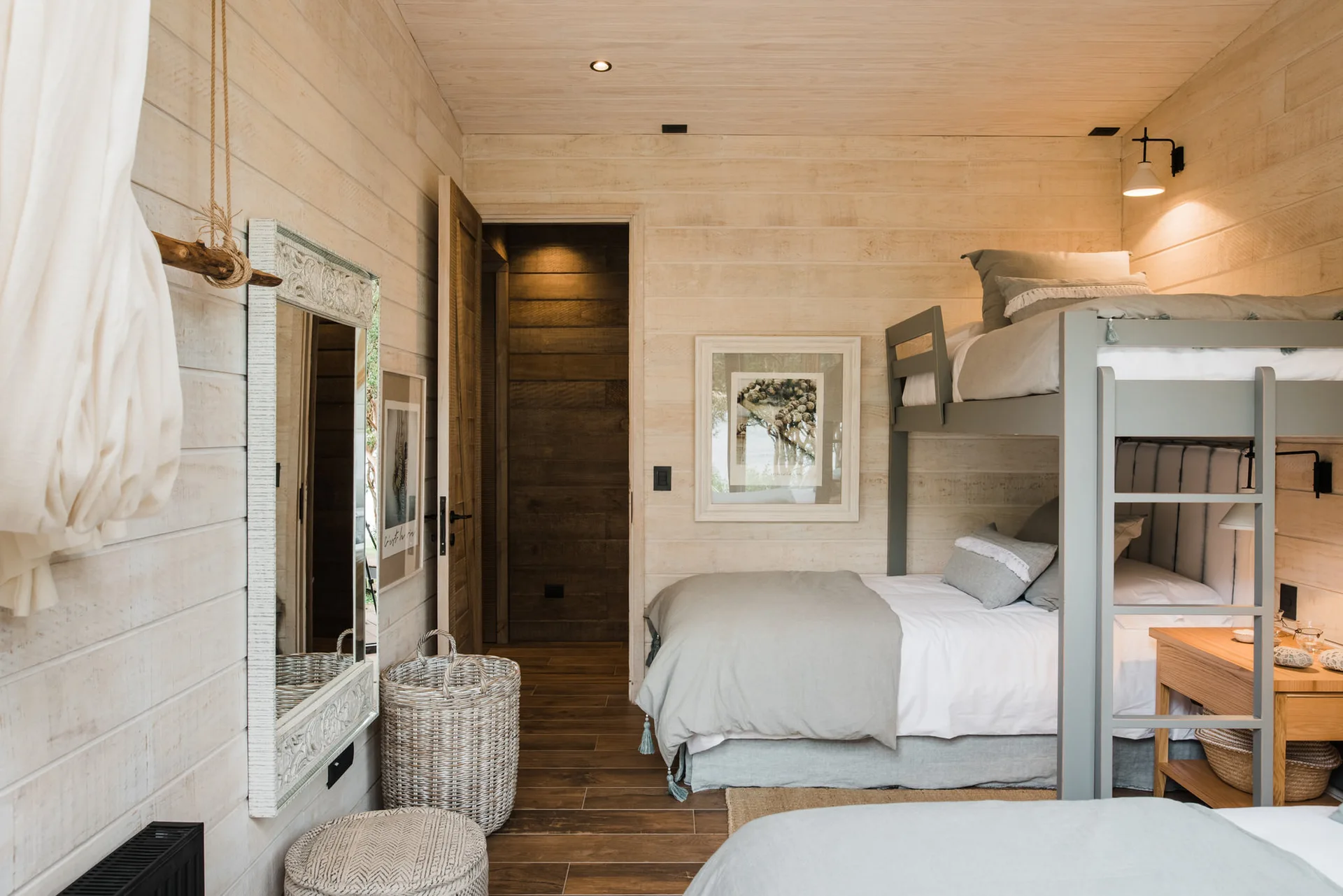 A rustic kids bedroom featuring light wood plank walls, a grey wooden bunk bed with muted bedding, a matching nightstand, and a decorative wall mirror.
