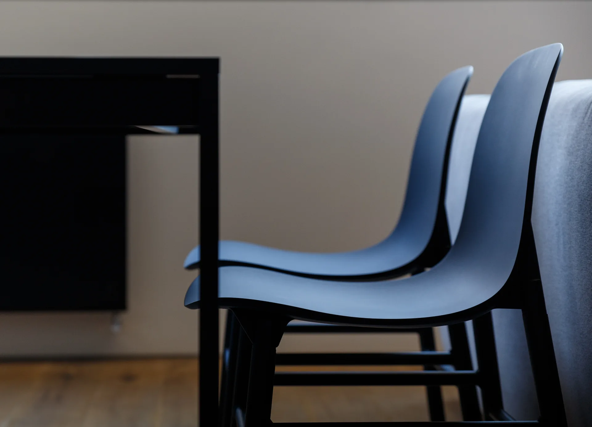 Two matte black molded dining chairs stand next to a black table on a wooden floor against a beige wall.