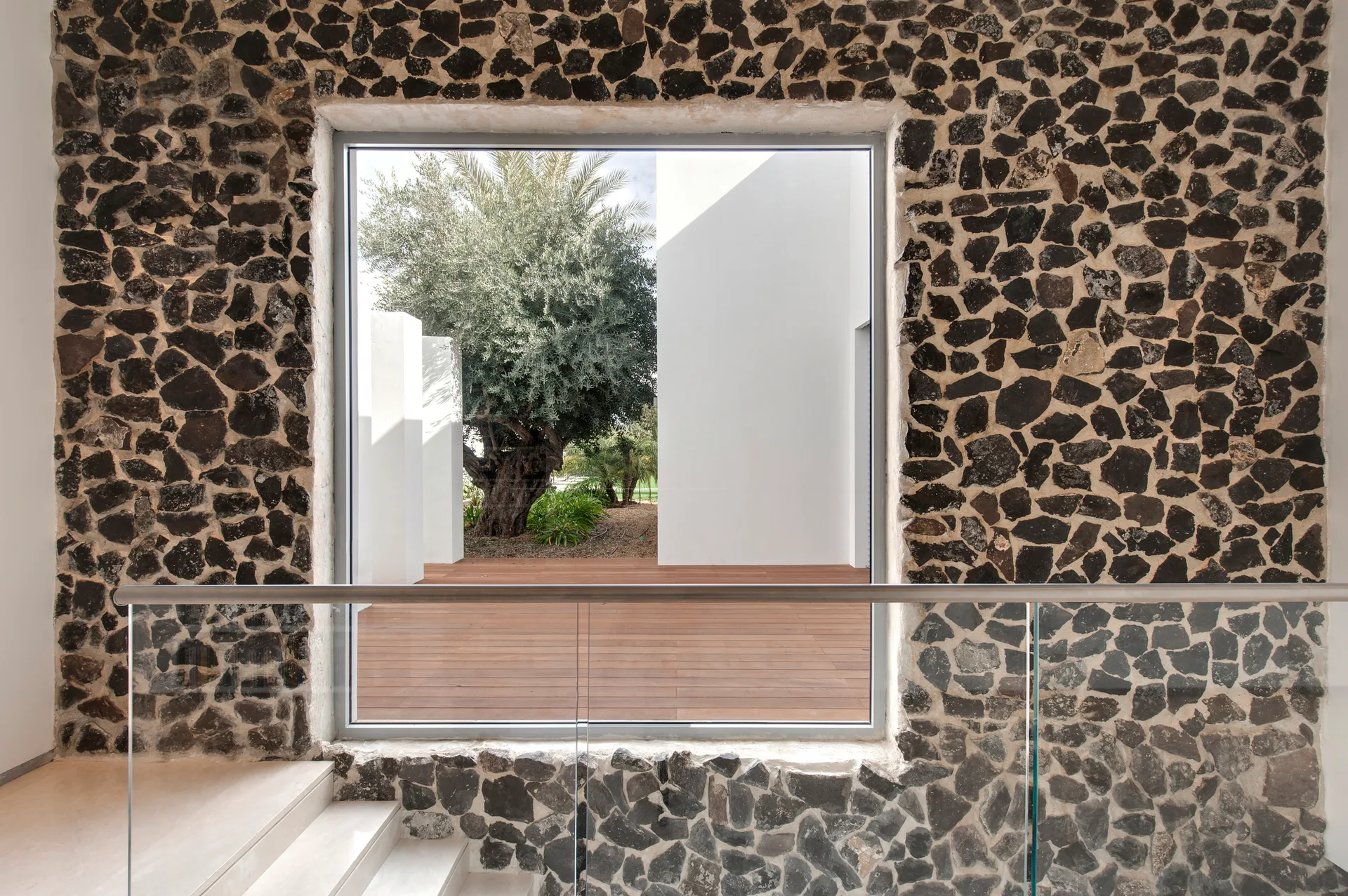 An interior staircase landing features a dark stone masonry wall, a glass railing, and a large window looking out at a courtyard.