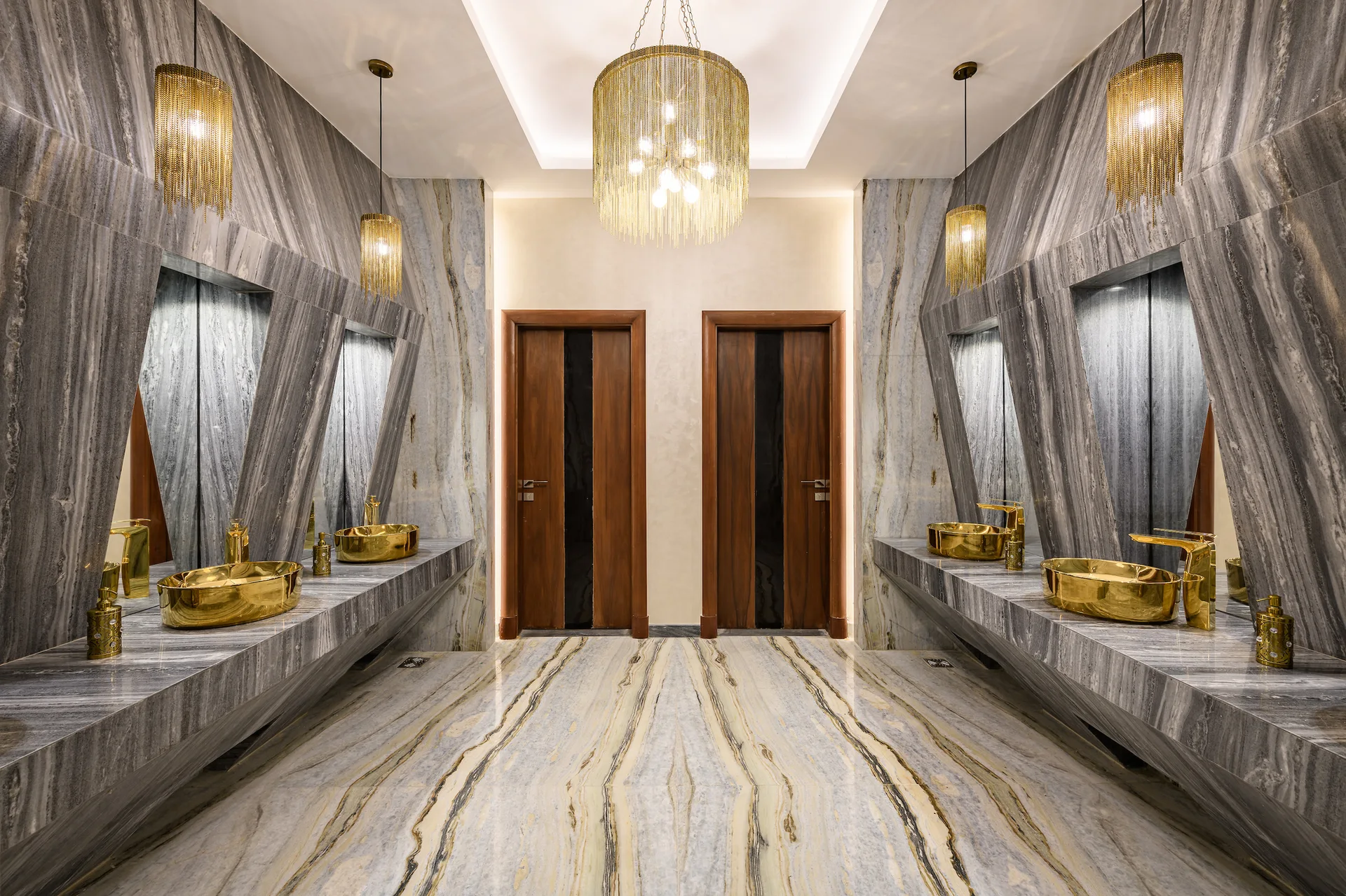A symmetrical bathroom featuring grey veined marble walls and floors, two floating vanities with gold vessel sinks, and fringed gold lighting.