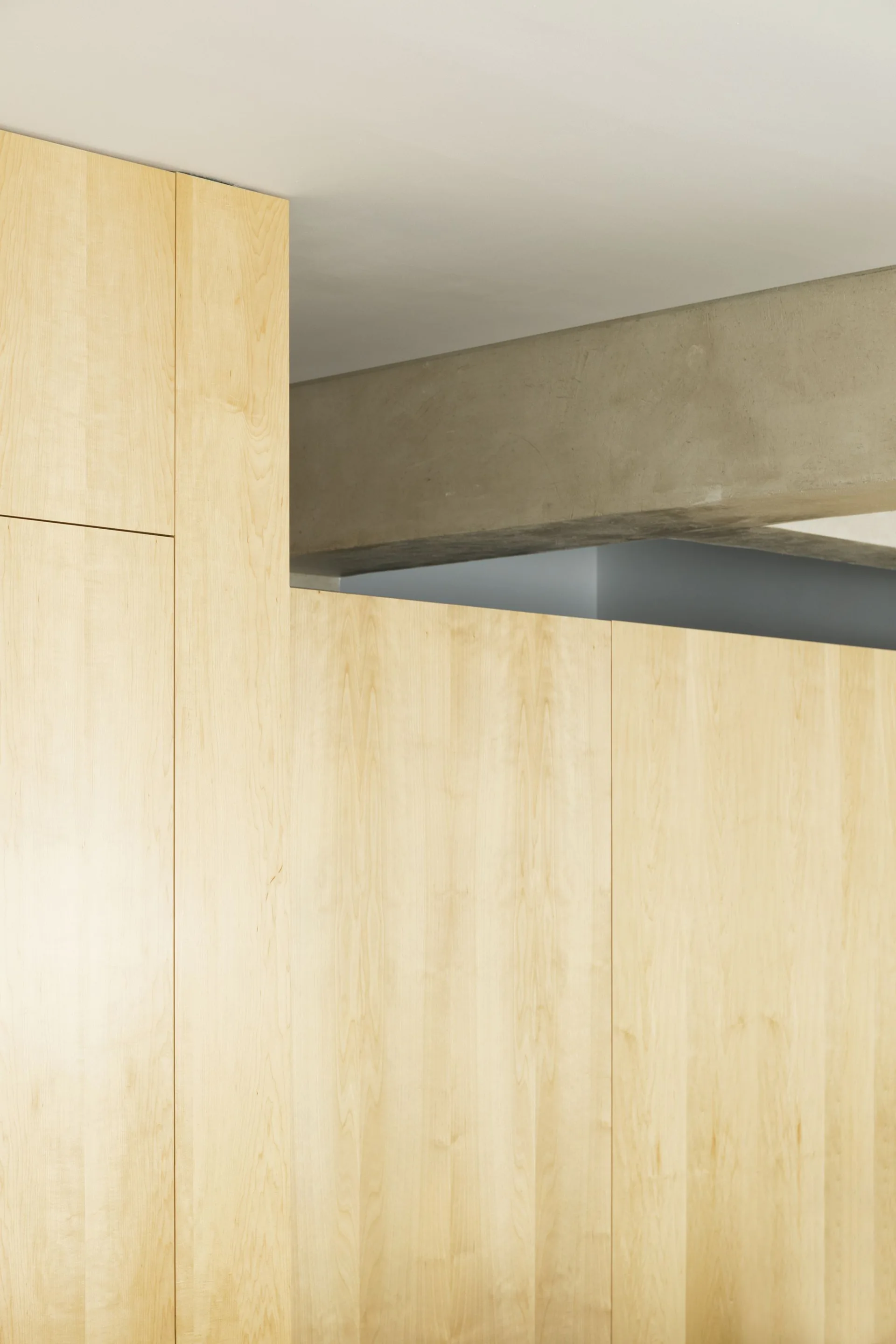 A close-up view of light wood cabinetry panels set against an exposed concrete structural beam and a gray ceiling.