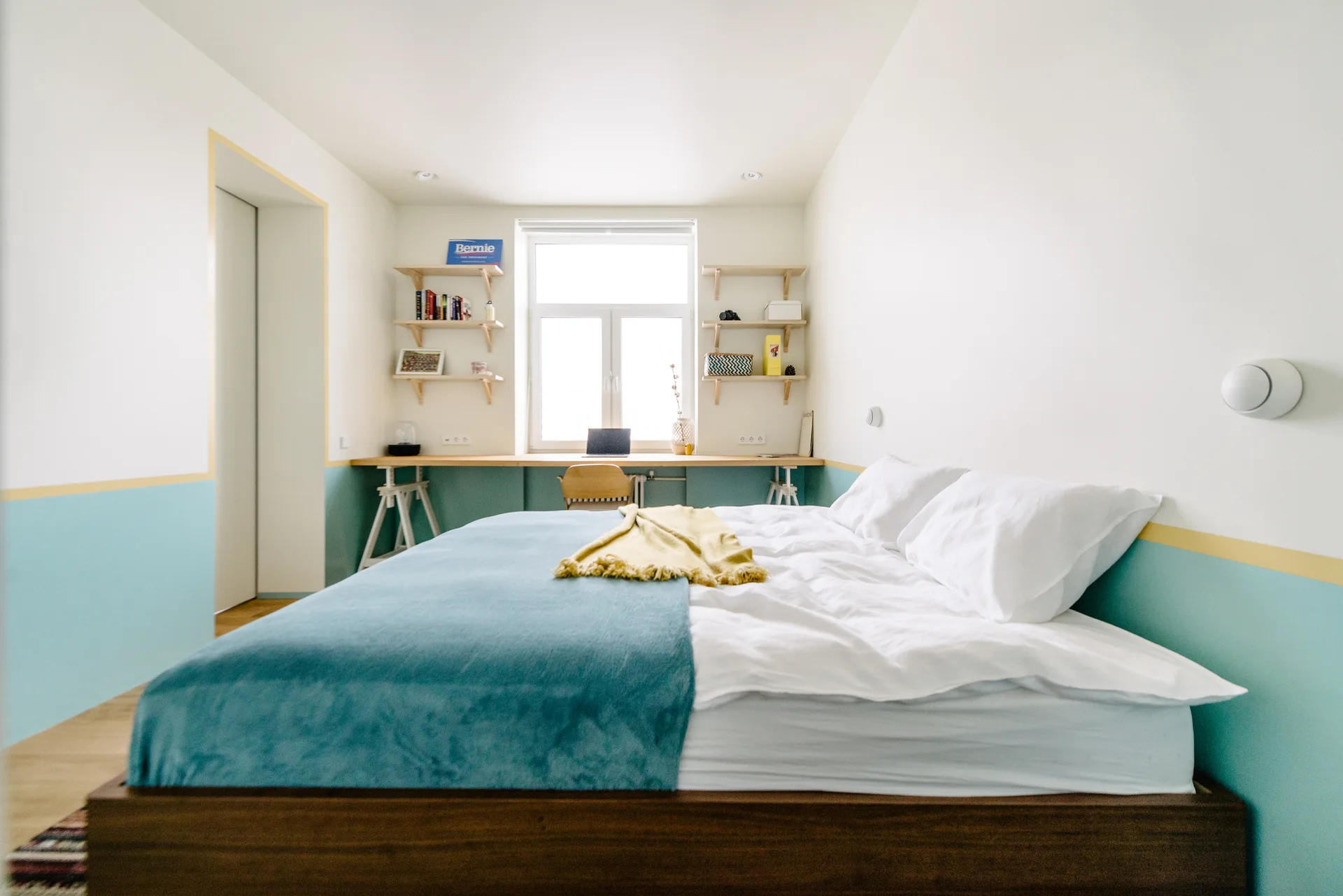 A minimalist bedroom features a dark wood bed frame, white and blue bedding, and a long built-in wooden desk positioned under a bright window.