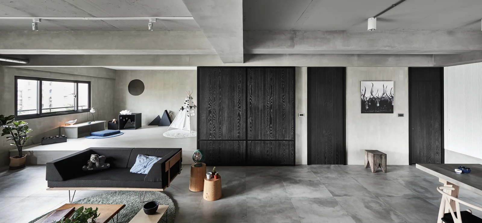 A minimalist industrial living room features raw concrete walls, a low black sofa, and a raised platform play area in the background.