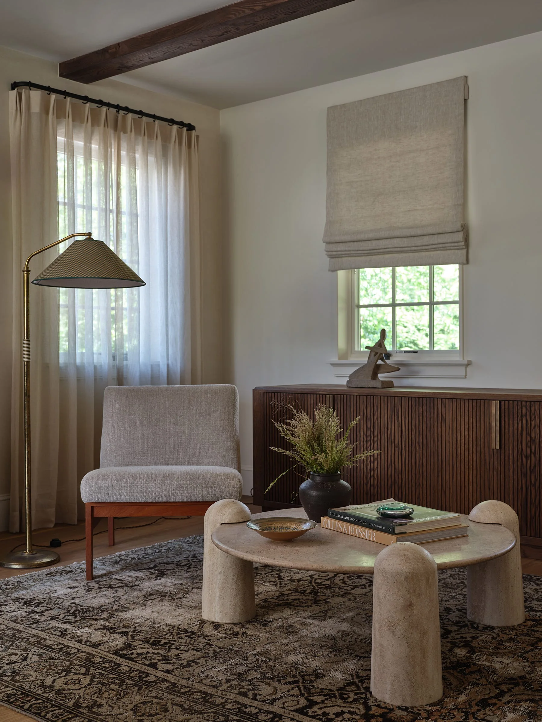 An upholstered armchair sits on a patterned rug next to a round travertine coffee table and a slatted wood credenza in a neutral living room.