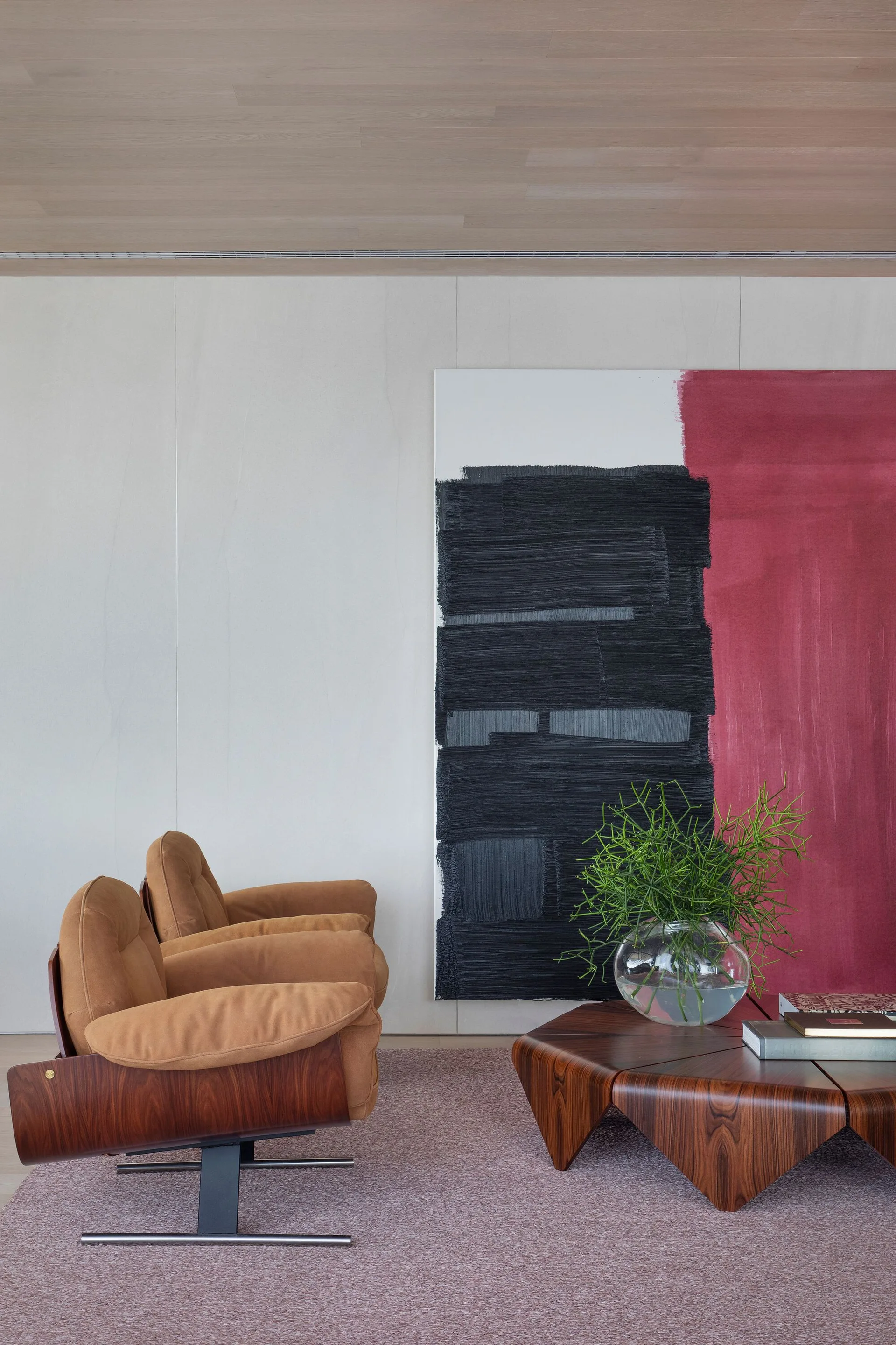 Two tan armchairs with curved wood frames sit on a mauve rug beside a sculptural coffee table and a large abstract painting.