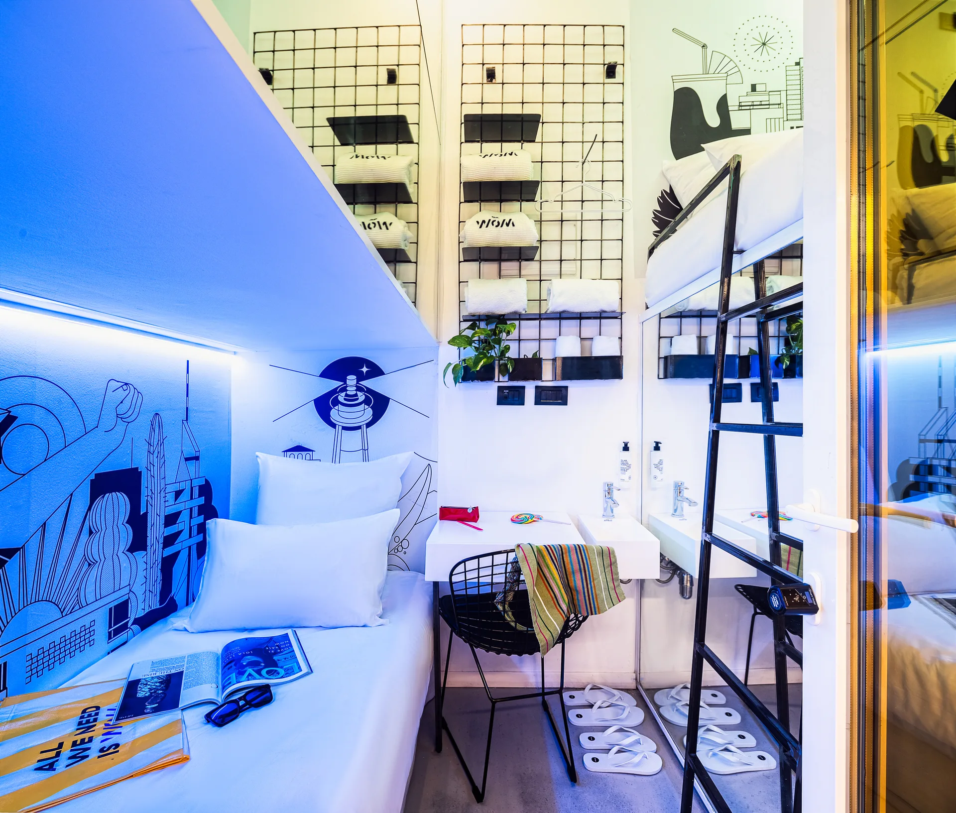 A compact micro-bedroom features white bunk beds, a black metal wire chair at a small desk, blue graphic mural art, and cool LED lighting.