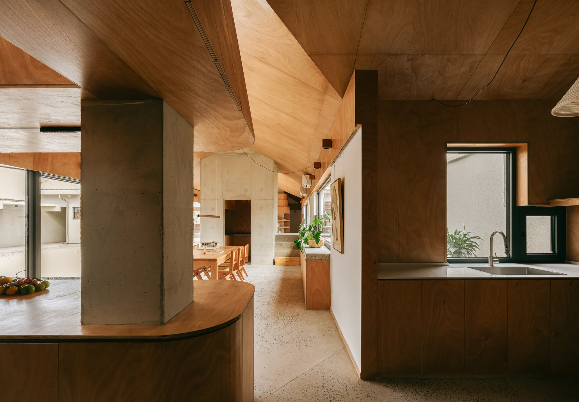A minimalist kitchen and dining area featuring warm plywood walls, an angular wooden ceiling, and an exposed raw concrete support column.
