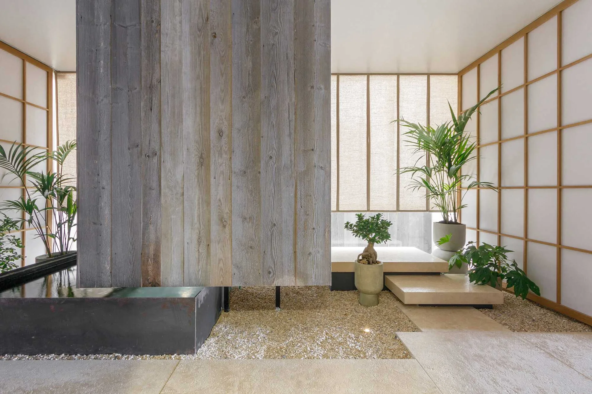 A minimalist indoor garden with a large weathered wood partition, a tranquil water basin, gravel flooring, and translucent shoji screens.