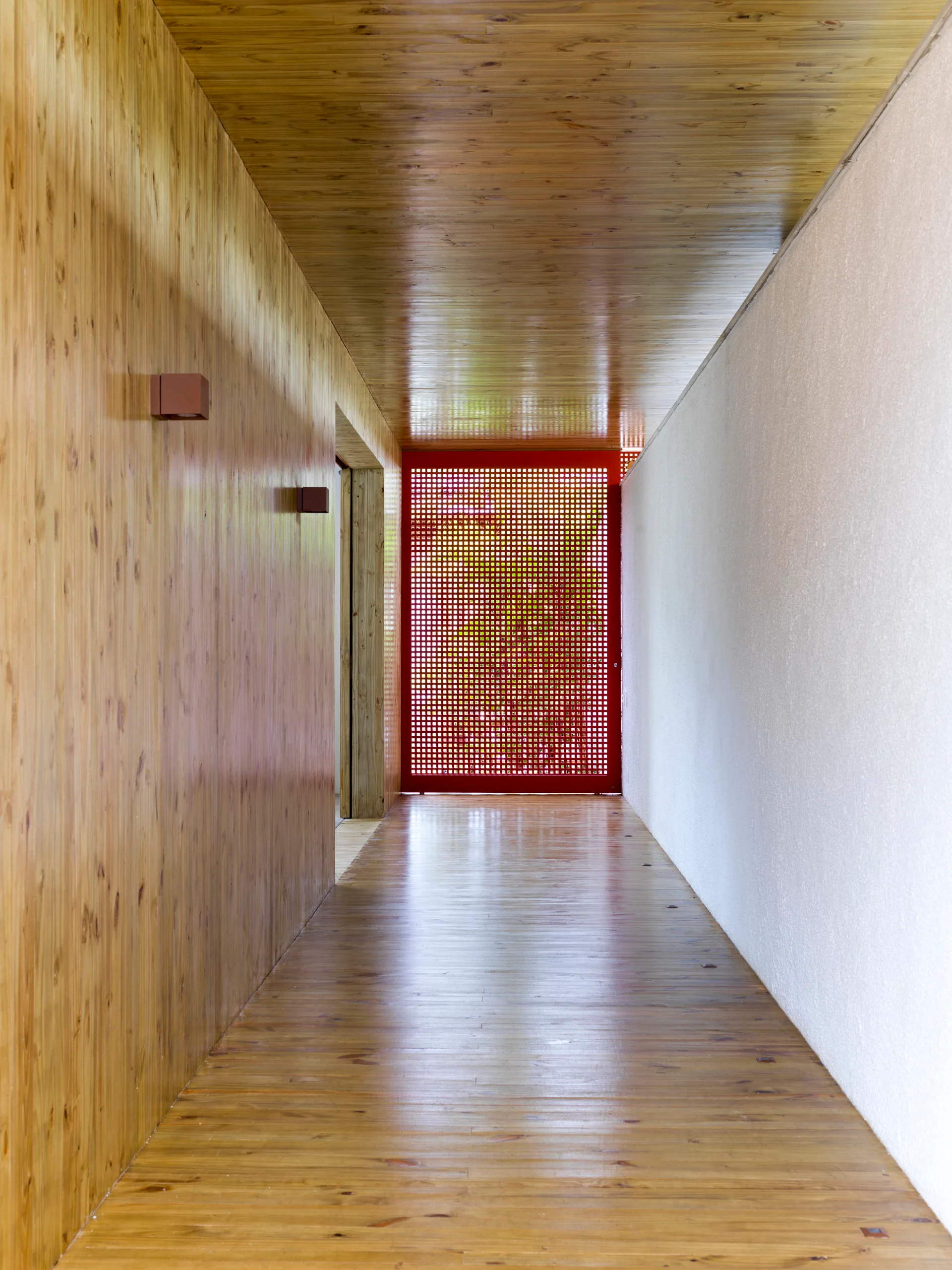 A long hallway with wood paneled walls, floor, and ceiling, a white wall on the right, and a bright red grid lattice screen at the end.