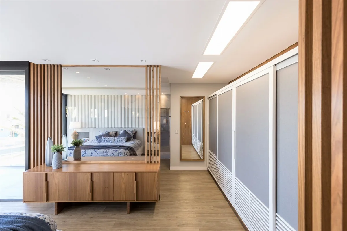A modern bedroom features a wooden slat room divider with an integrated mirror reflecting the bed next to a large sliding wardrobe.