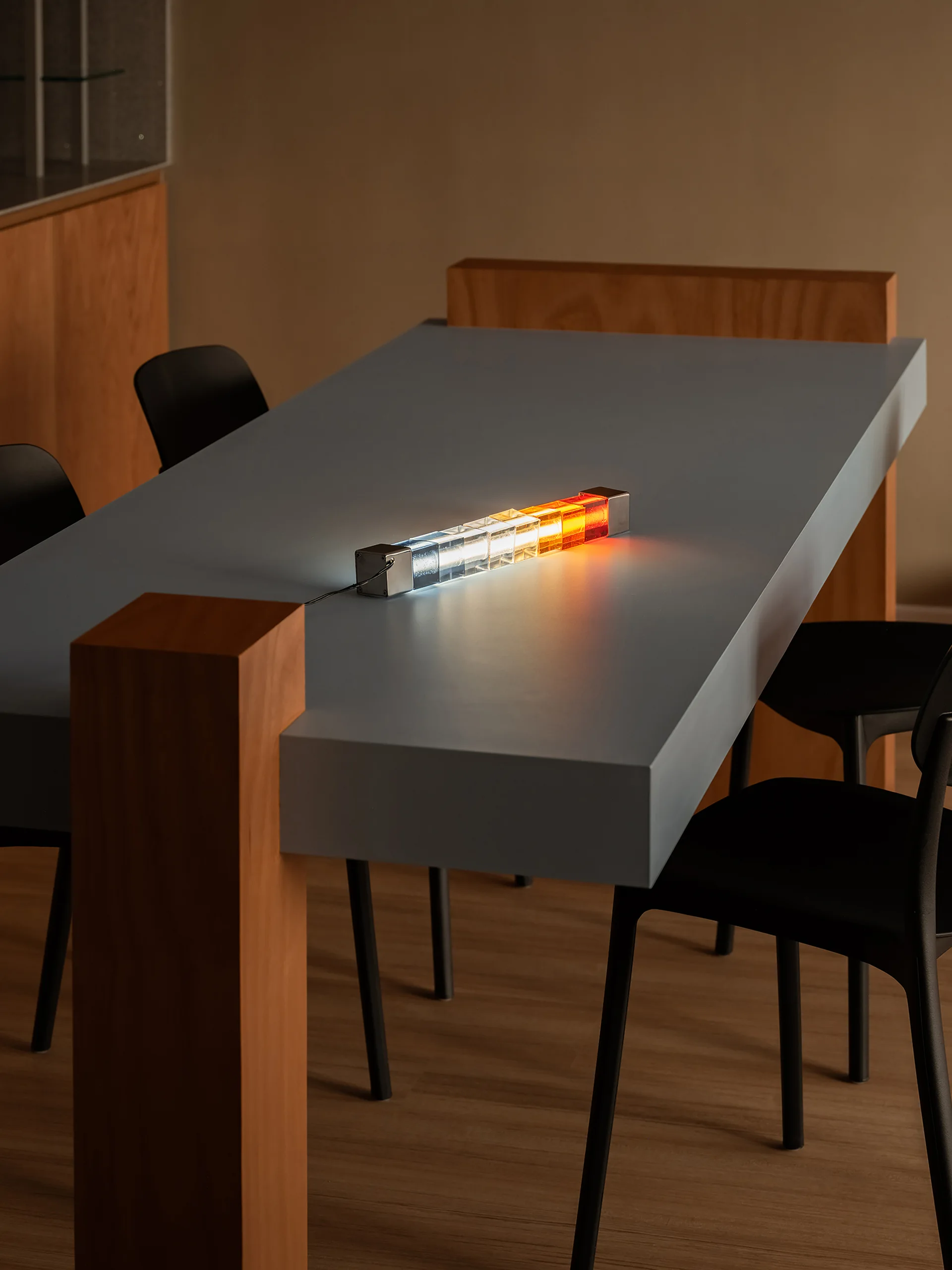 A minimalist blue dining table with thick wood legs and black chairs features a glowing multi-colored geometric table lamp.