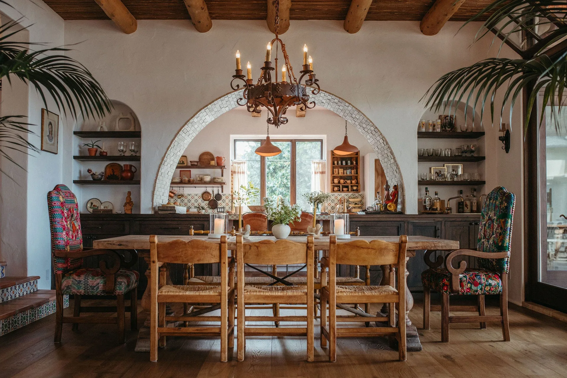 A wooden dining table sits beneath a wrought iron chandelier and exposed log ceiling beams, with a tiled archway in the background.