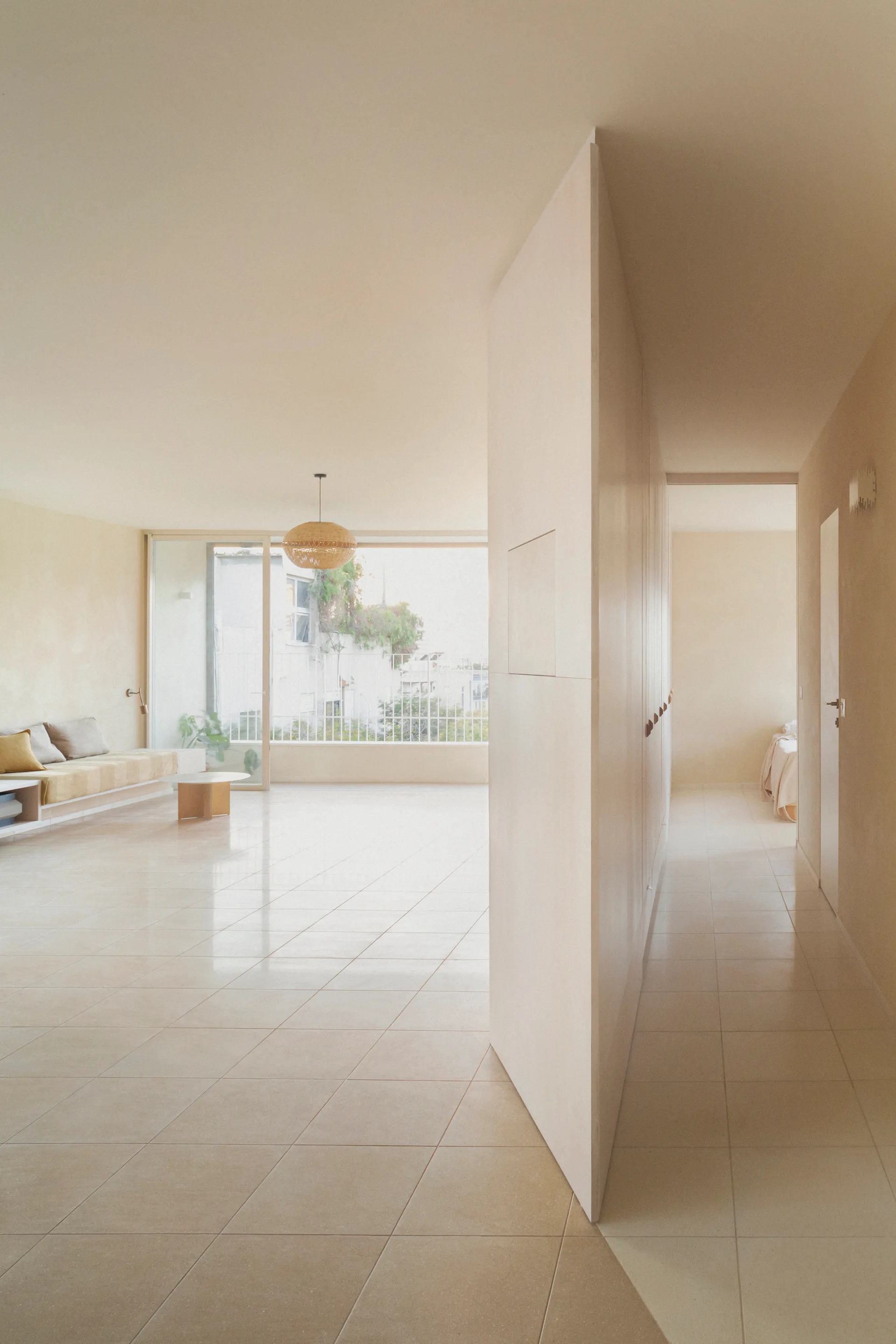 A minimalist beige living room featuring a central partition wall, tiled flooring, a low-profile sofa, and a hallway leading to a bedroom.
