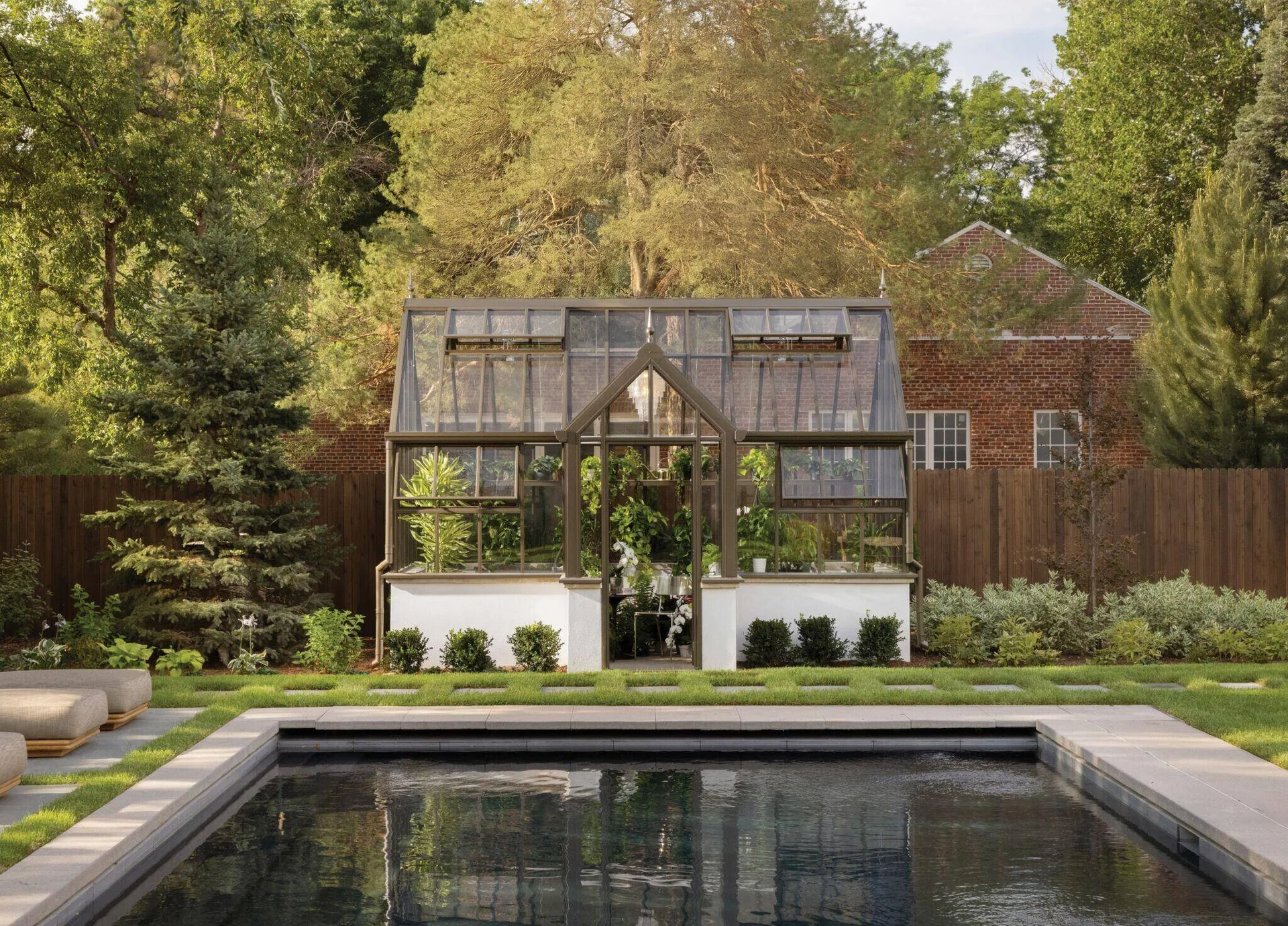 A traditional glass greenhouse with a white stucco base stands behind a rectangular swimming pool in a lush landscaped backyard.