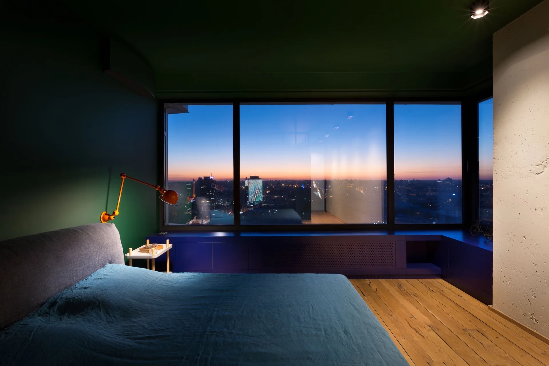 A modern bedroom features deep green walls, a bed with blue linens, and a low blue storage bench below a large window showing a city sunset.