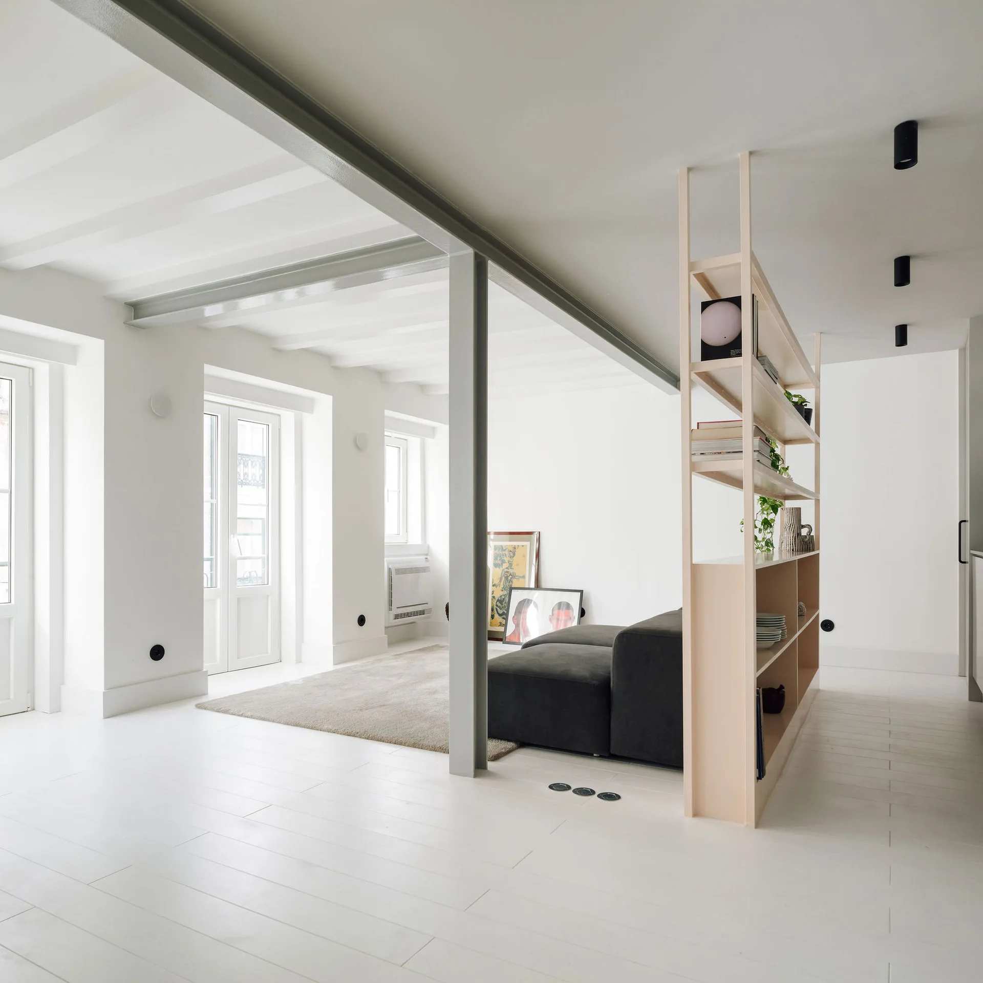 A minimalist living room with white floors, grey structural beams, a dark sectional sofa, and a light wood open-shelving partition.