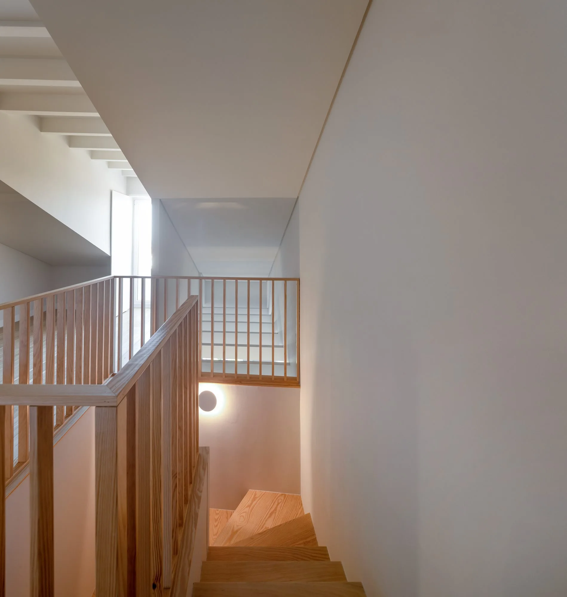 A light wood staircase with vertical slat railings leads down a minimalist stairwell with plain white walls and a glowing round wall sconce.
