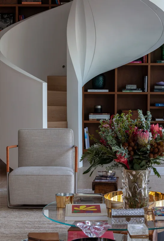 A contemporary living space features a curved white spiral staircase next to a wooden bookshelf, with an upholstered armchair and glass coffee table in front.