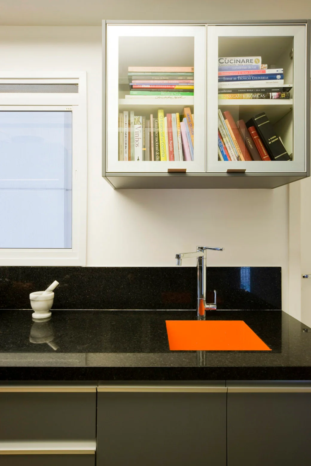 A modern kitchen details a black granite countertop with a bright orange sink basin and glass-front upper cabinets filled with books.