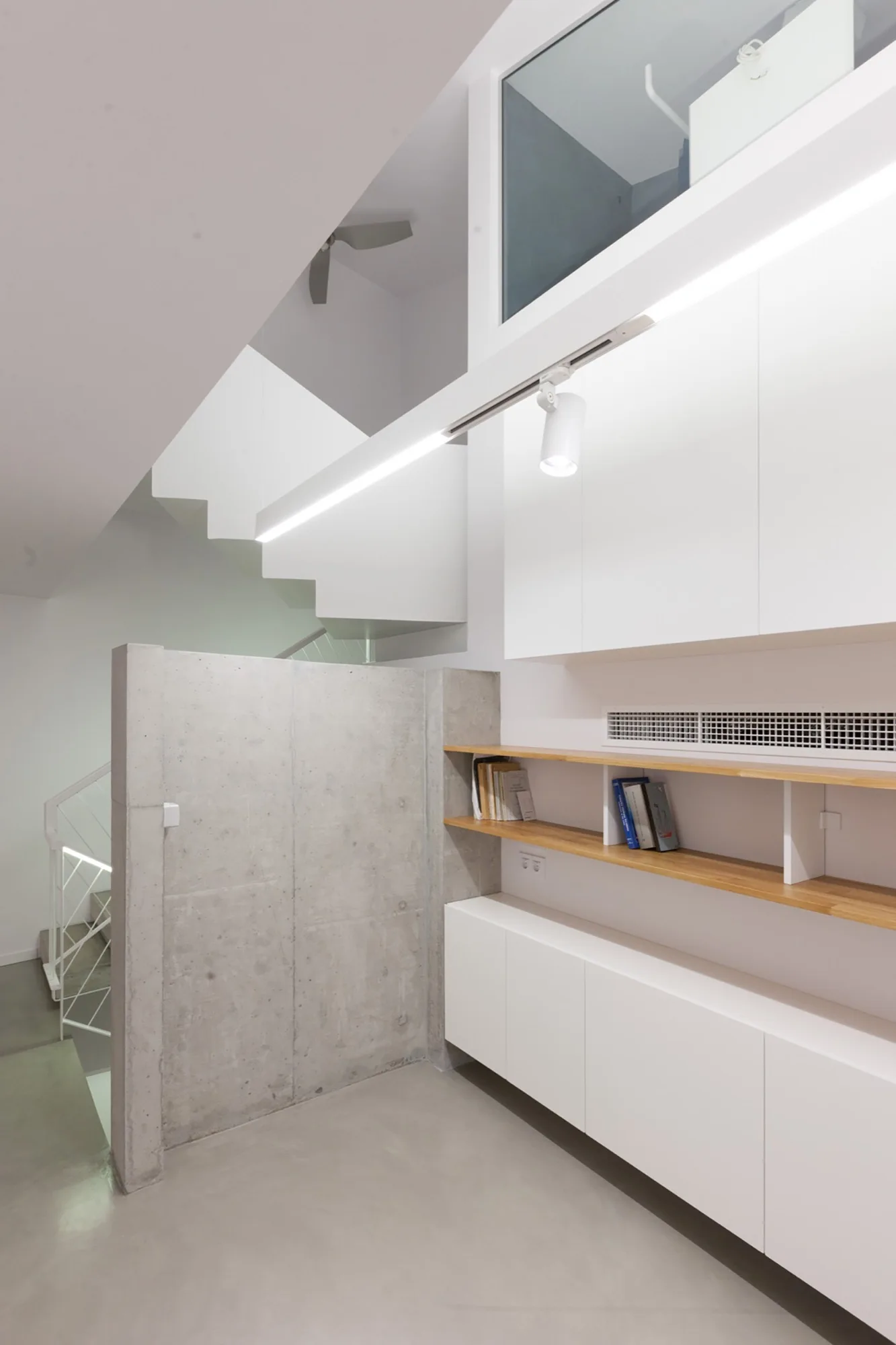 A modern interior space features an angular white staircase, a smooth concrete partition wall, and white built-in cabinetry with natural wood shelving.