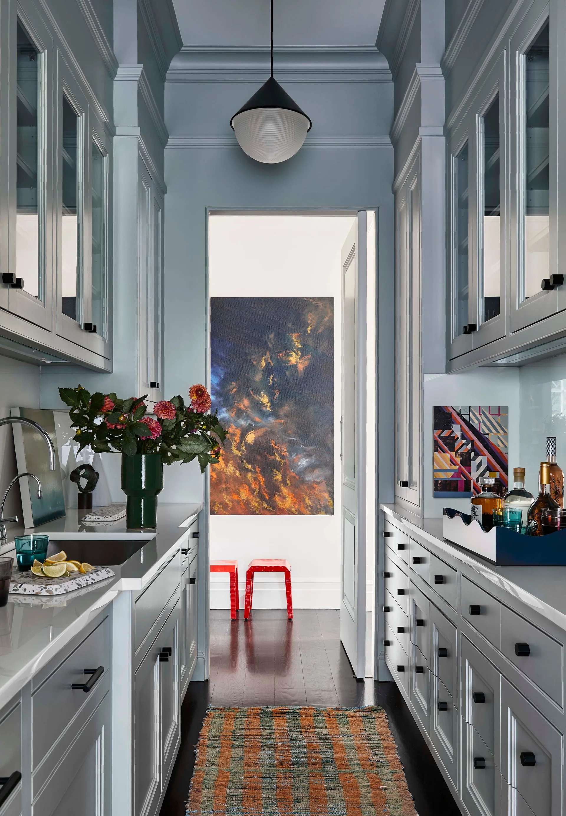 A galley-style butler's pantry features pale blue cabinetry, white countertops, glass-front upper cabinets, and a doorway opening to a room with colorful art.