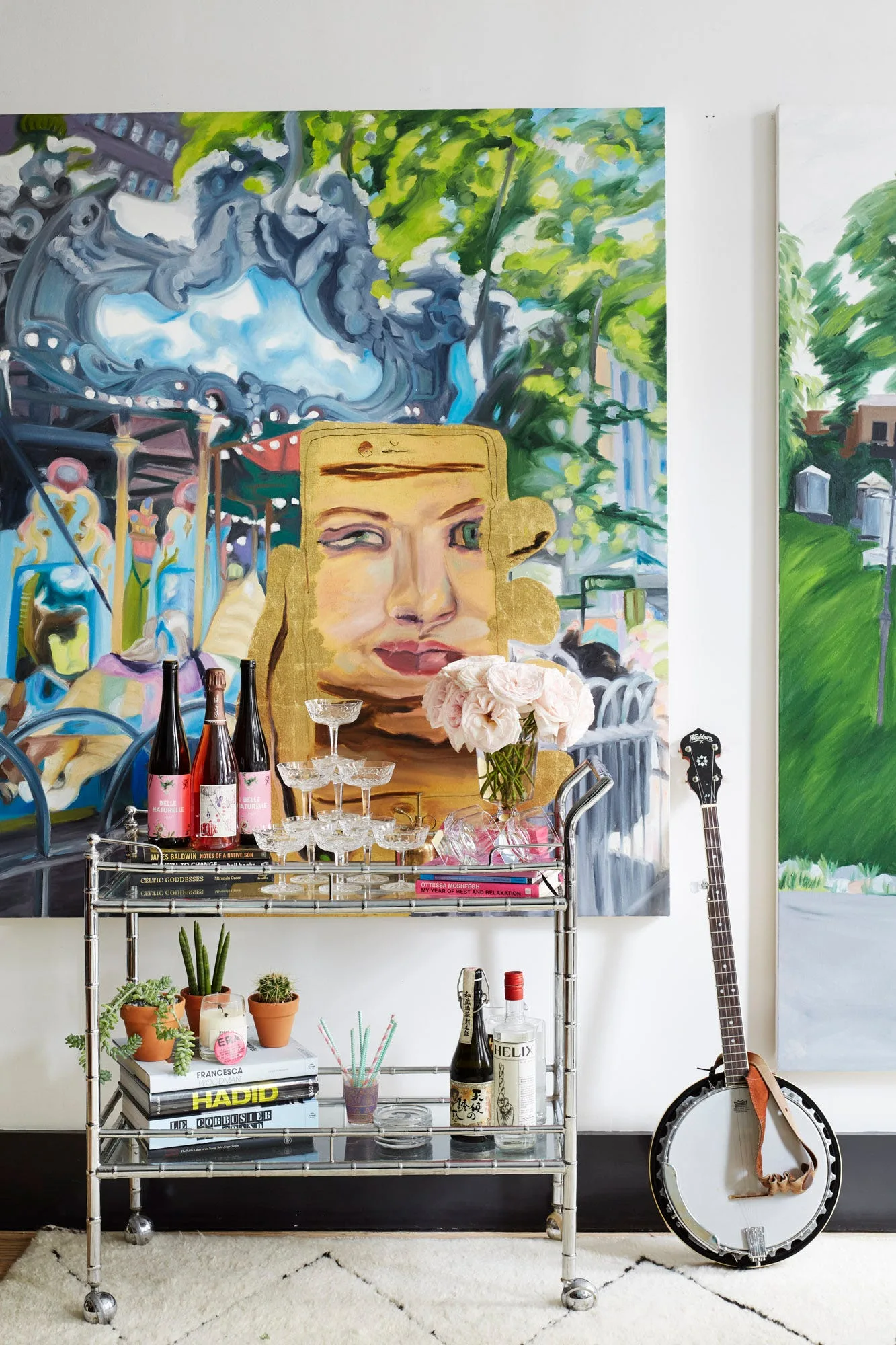 A chrome two-tiered bar cart styled with glasses, bottles, and flowers sits in front of two large colorful paintings, with a banjo leaning nearby.