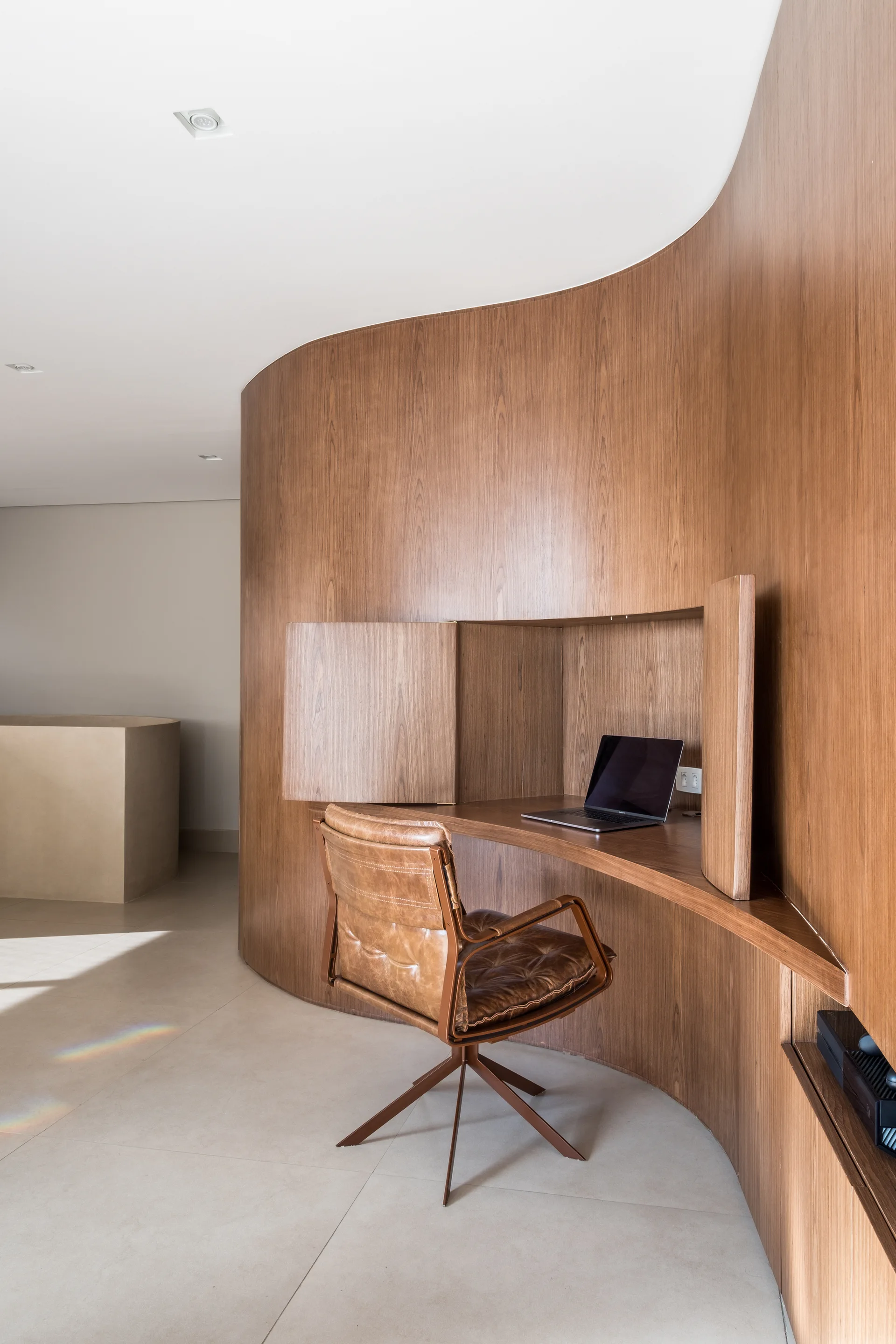A home office featuring a curved wood paneled wall with a built-in desk nook, a laptop, and a brown leather swivel chair.