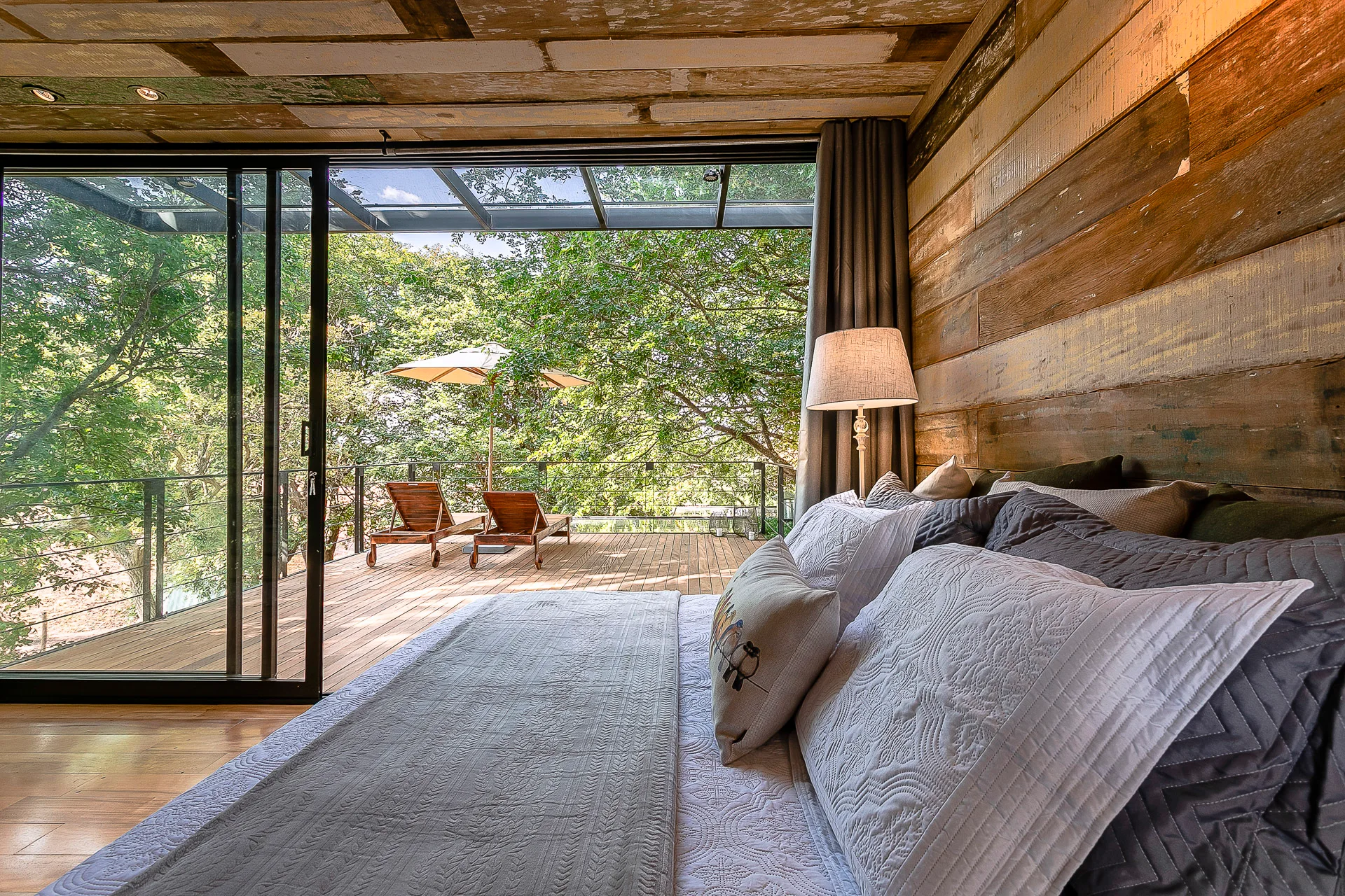 A rustic bedroom features layered grey bedding, a weathered wood wall, and sliding glass doors that open to a wooden deck with lounge chairs.